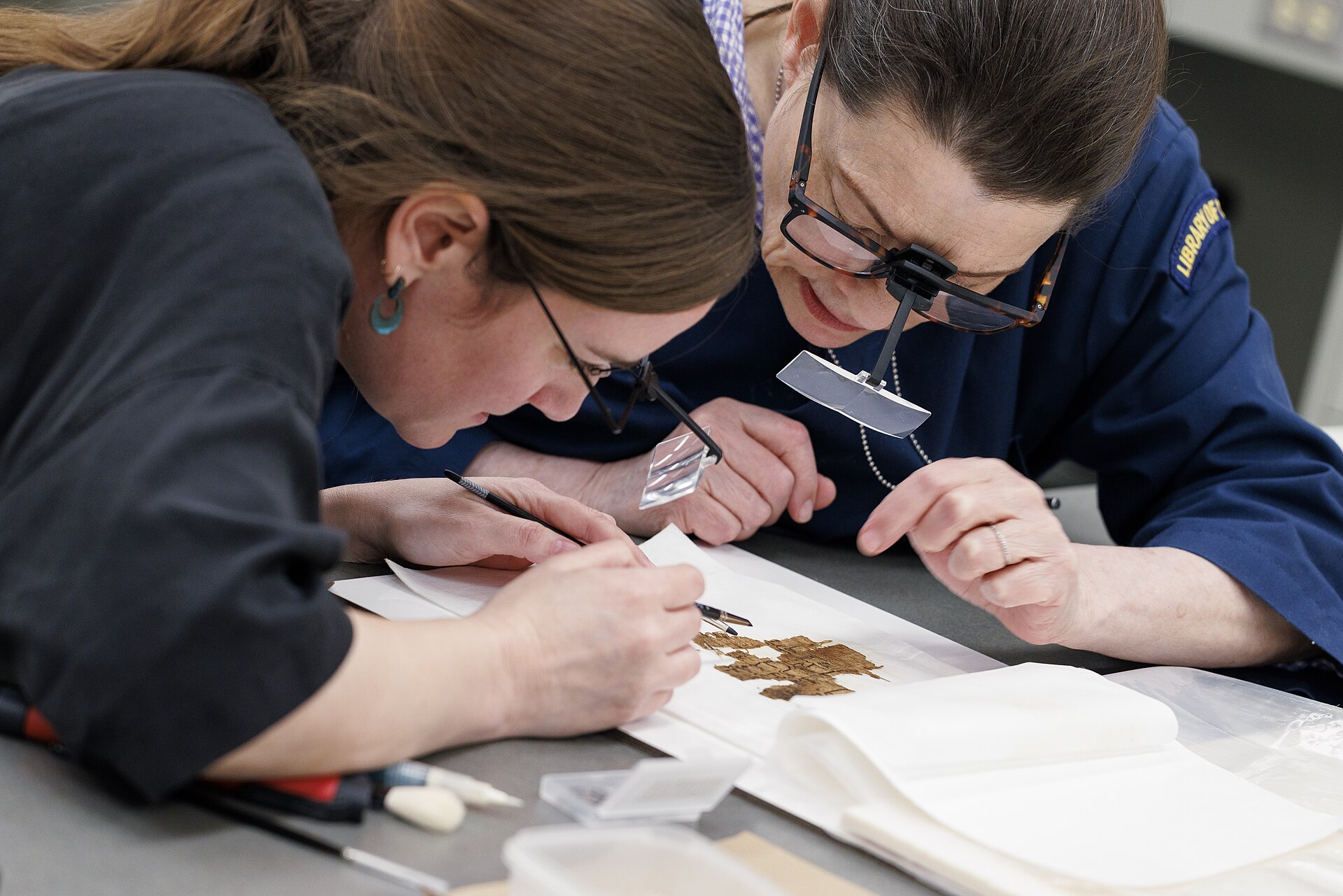 Gwenanne Edwards, Senior Paper Conservator in the Conservation Division of the Library of Congress, and her colleague Claire Dekle treating a papyrus fragment from the Fustat Collection of the African and Middle Eastern Division on 25 April 2025; photograph by Shawn Miller for the Library of Congress. The collection is more than fifty administrative texts from Egypt of about the seventh to eleventh centuries CE, mostly Arabic with a few in Greek and Coptic, recovered archaeologically from a midden at Fustat, now part of Cairo, and they reached the bench crumpled, folded and covered in debris with the fibres lying askew across the writing. What is happening here is the whole of the treatment: humidity is introduced to make brittle papyrus flexible, then the fibres and the detached fragments are unfolded, aligned and reattached one at a time under a magnifier. Edwards learned papyrus conservation only in 2024, at the University of Michigan Library's Papyrus Conservation Seminar, and afterwards prepared and taught the technique to her own division, which treated about half the collection in the workshop that followed.
