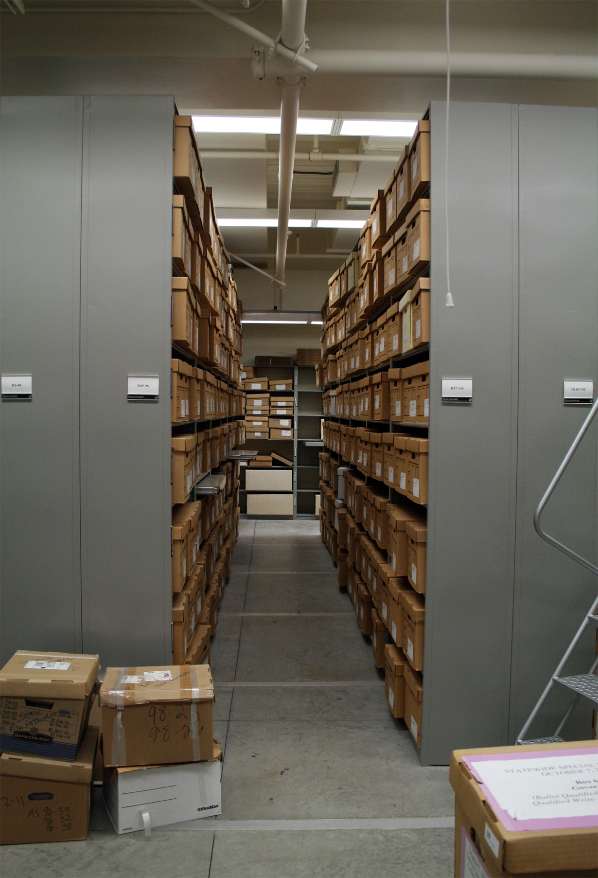 Document boxes on mobile shelving in the California State Archives, Sacramento. Each shelf range carries its own reference label, and boxes awaiting shelving are stacked on the floor with their numbers written on the lid.