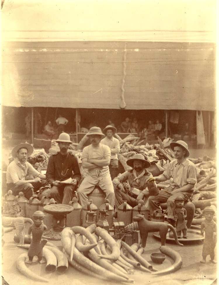 Six European members of the British punitive expedition seated among ivories and cast brasses taken from the royal palace at Benin City, photographed 18 February 1897.