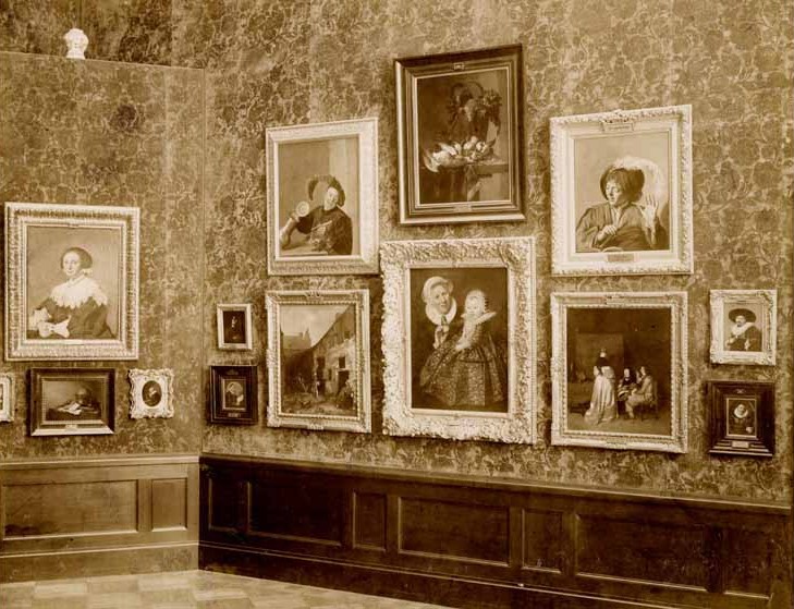 Room 59 of the Kaiser-Friedrich-Museum in Berlin, photographed in 1904: seventeenth-century Dutch pictures hung in tiers in Wilhelm von Bode's arrangement, several frames carrying their own small name-plates. At the upper left is The Merry Drinker, canvas, about 1629, 77 by 63 cm, Gemaeldegalerie, Staatliche Museen zu Berlin, inventory number 801B. It came into the collection in 1874 with the purchase of the Suermondt collection at Aachen, was first given to Frans Hals, and holds an inventory number inside the museum's Frans Hals block, yet Bode's own printed catalogue of 1904, in force when this photograph was taken, already enters it under Judith Leyster; Leyster painted a second version of the subject, now in the Rijksmuseum, Amsterdam, so the Berlin picture is identified here by its accession number. The label had already moved by 1904 and the accession number never did.
