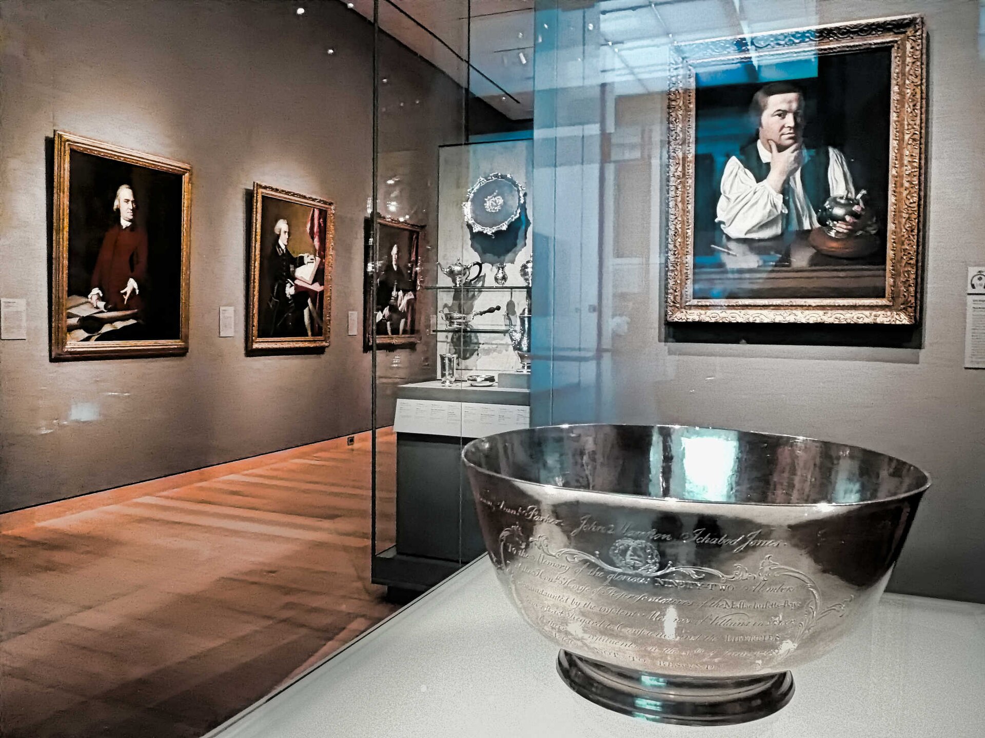 The Sons of Liberty bowl on display at the Museum of Fine Arts, Boston, accession 49.45, with Copley's portrait of Paul Revere on the wall behind it. The engraved names of the fifteen subscribers run below the rim. The Museum's public record gives the bowl's dimensions and no weight.