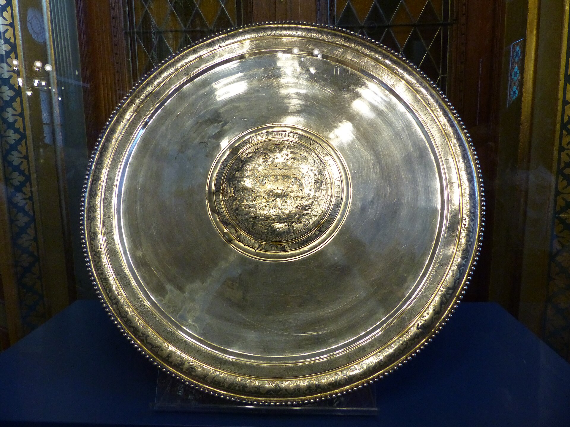 The Hunting Plate of the Sevso Treasure, 70.5 cm across and 8.8 kg of silver, photographed in the display of the Hungarian National Museum. The central medallion holds an open-air banquet ringed by hunting scenes, and around it runs the hexameter couplet that gives the treasure its name, together with the lake labelled PELSO that is read as Balaton. This photograph could only be taken because Hungary bought the silver. Before 2014 the plate's entire recorded viewing history was a Zurich bank vault, a museum inspection on approval and one invitation-only afternoon at Bonhams.