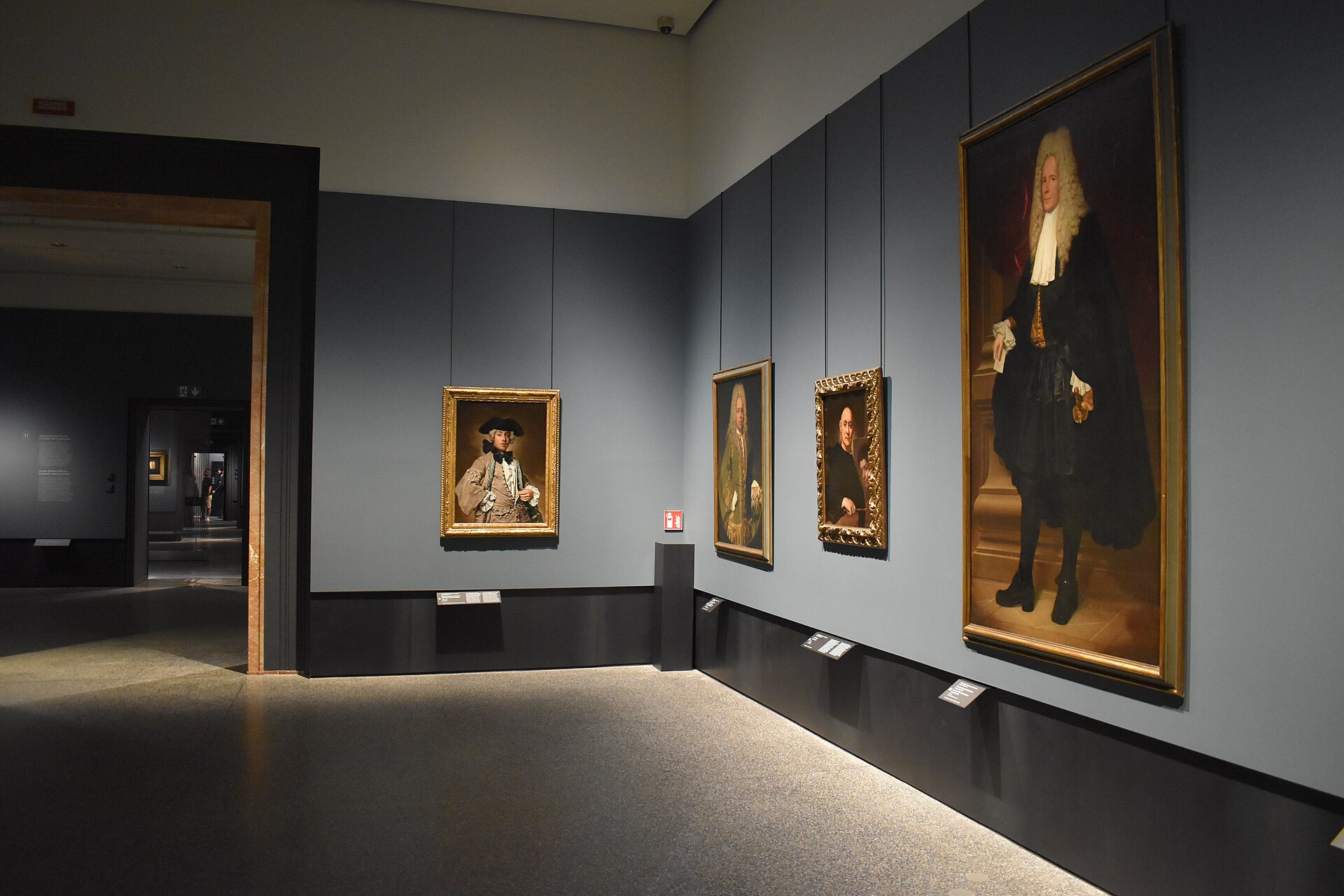 A grey-walled gallery hung with formal eighteenth-century portraits, including a full-length figure in a white wig and black robe