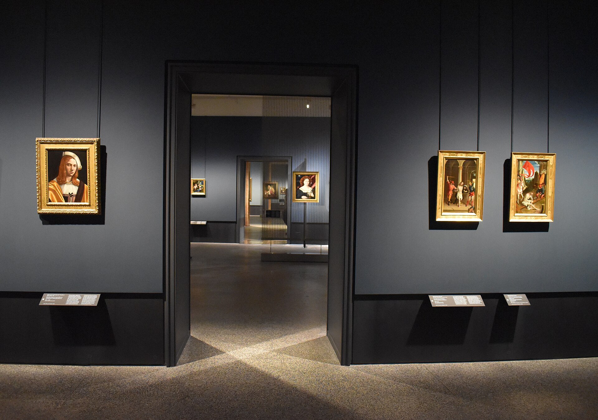 A dark-walled gallery hung with small gilt-framed early Renaissance panels, seen through an open doorway into a further room of the same sequence