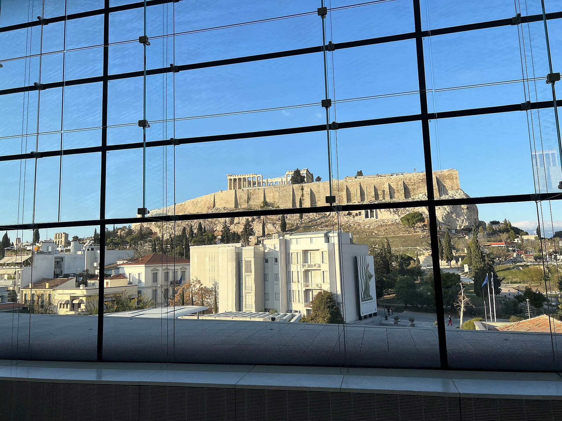 Looking through the grid of the Parthenon Gallery's glass wall toward the Acropolis and the Parthenon on the hill beyond