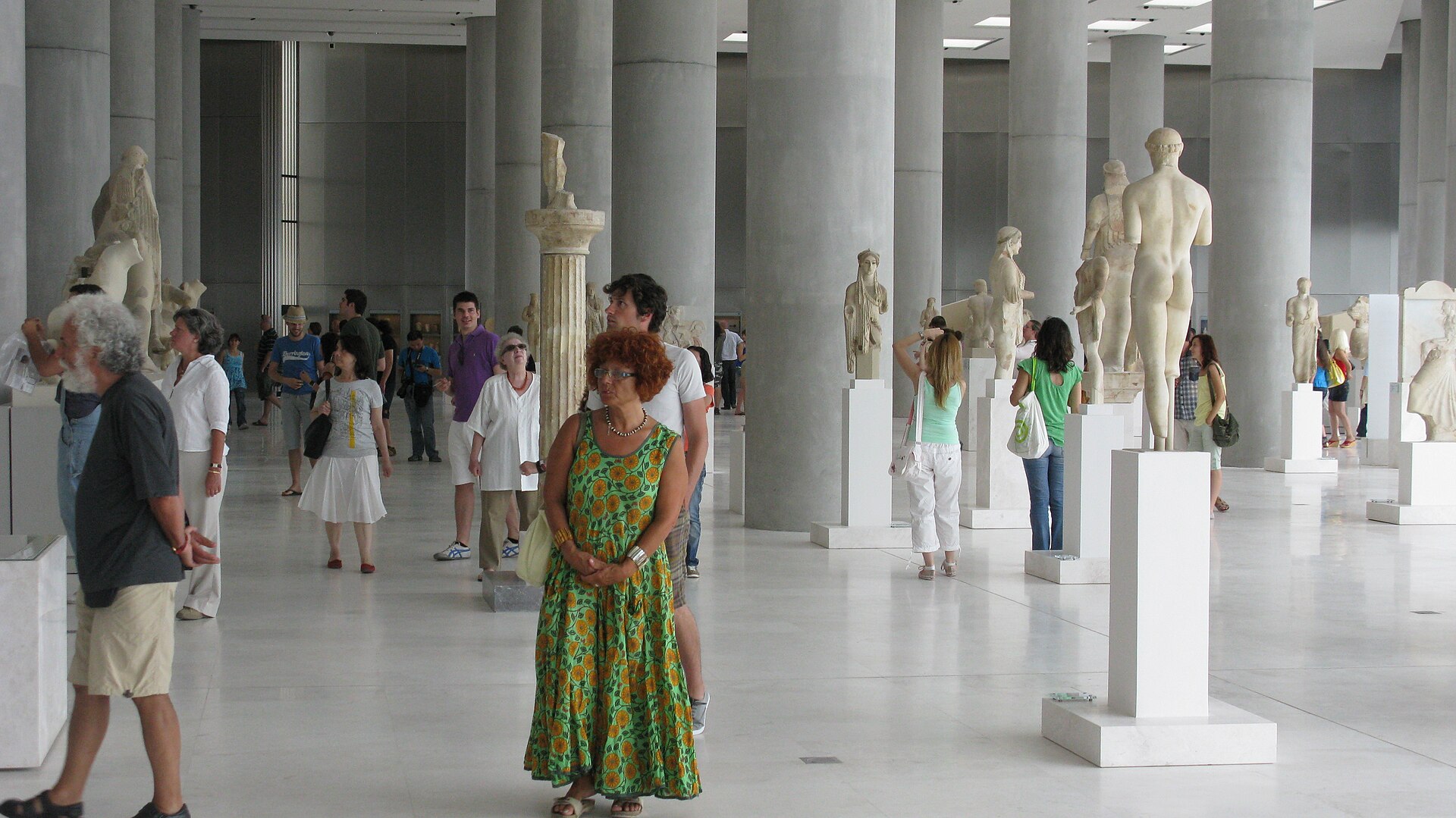 A colonnaded gallery with archaic marble statues, including standing draped figures, arranged between rows of columns
