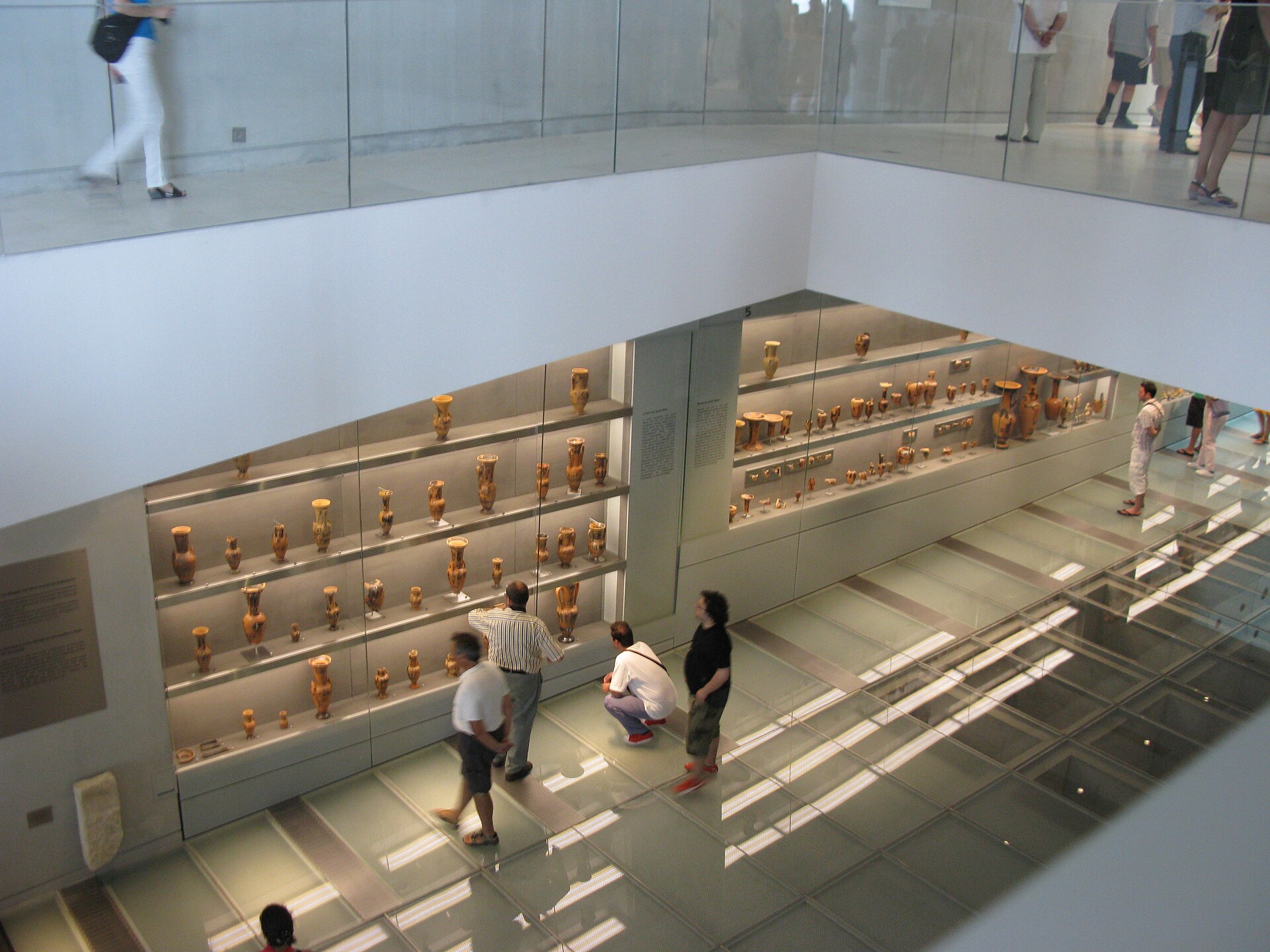 Illuminated wall cases of small vessels along the glass-floored ramp of the ground floor, visitors looking through the floor at the excavation below