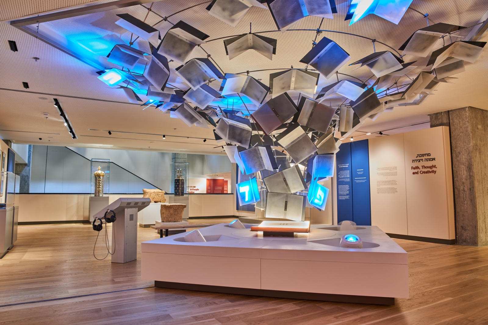 A suspended sculptural installation of angled panels bearing Hebrew letters over a low white plinth, with museum text panels reading Faith, Thought and Creativity in the background