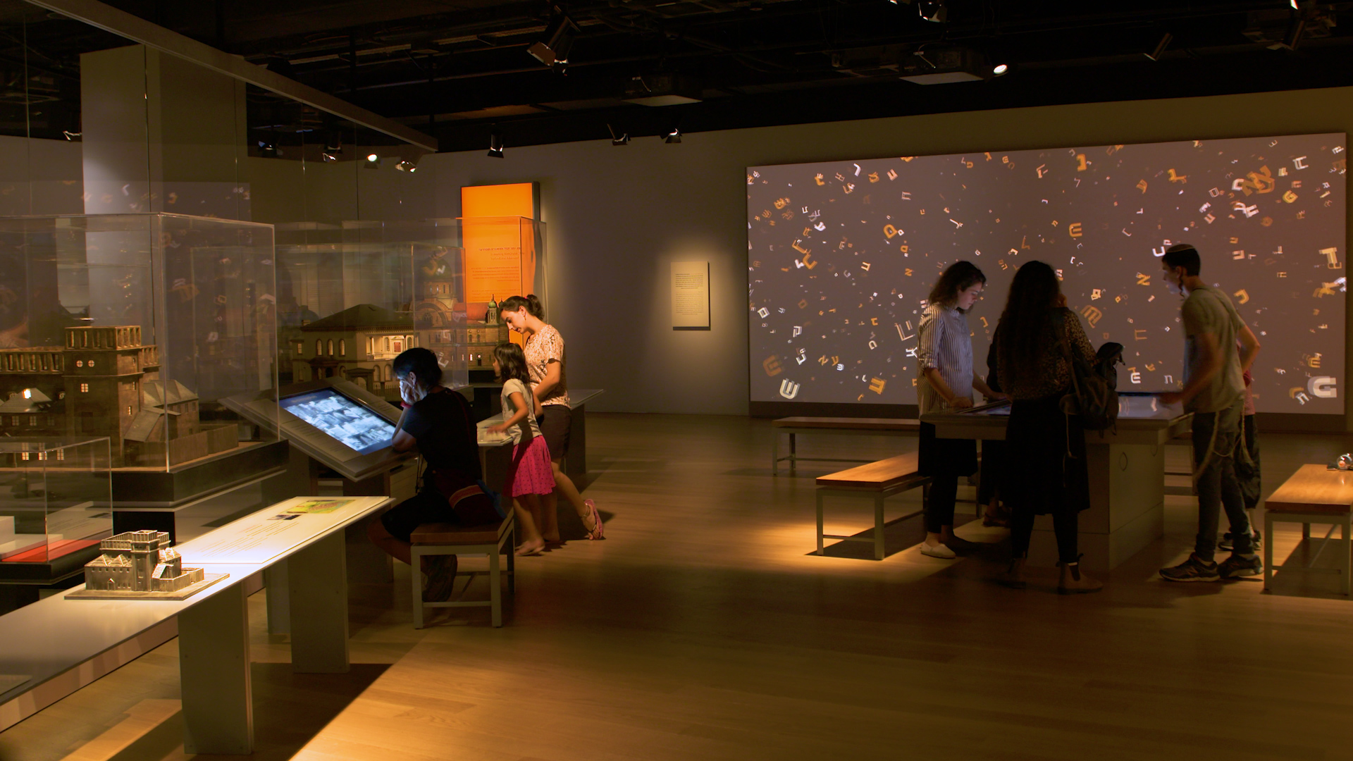 Glass display cases holding architectural models of historic synagogue buildings, with an interactive touchscreen table and an illuminated wall of falling Hebrew letters beyond