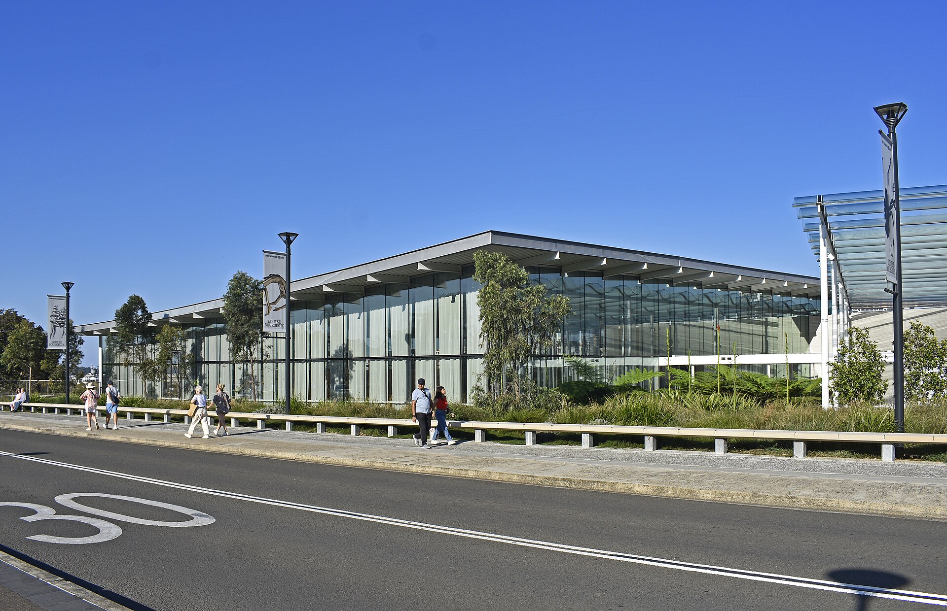 The glazed street facade of Naala Badu seen from Art Gallery Road, with native landscaping and an exhibition banner in front