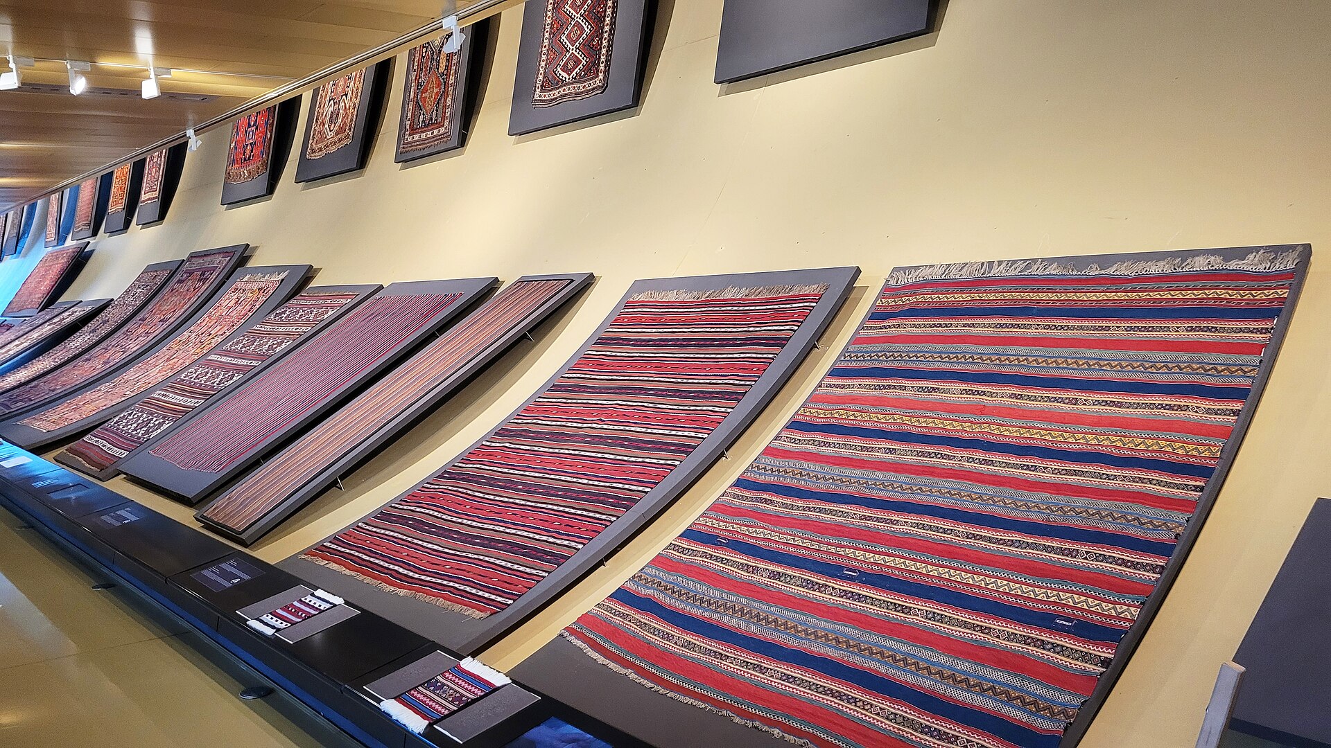 Rows of flat-woven Azerbaijani carpets and kilims mounted at an angle along a gallery wall