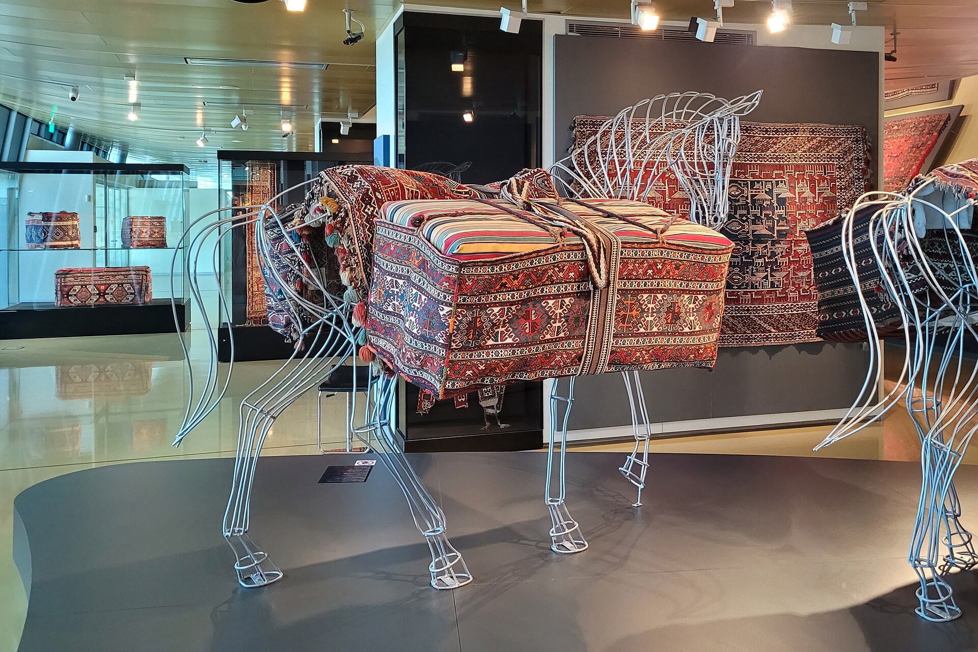 Wireframe model horses draped in woven Azerbaijani saddlebags and carpet trappings, with pile carpets on the wall behind