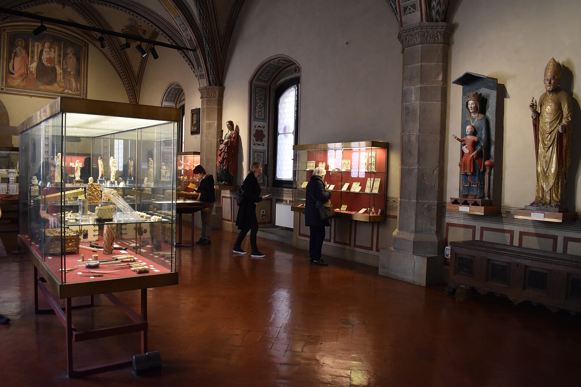 A gallery of ivory carvings and painted altarpieces in glass display cases, with a large gilded wood saint statue on the right