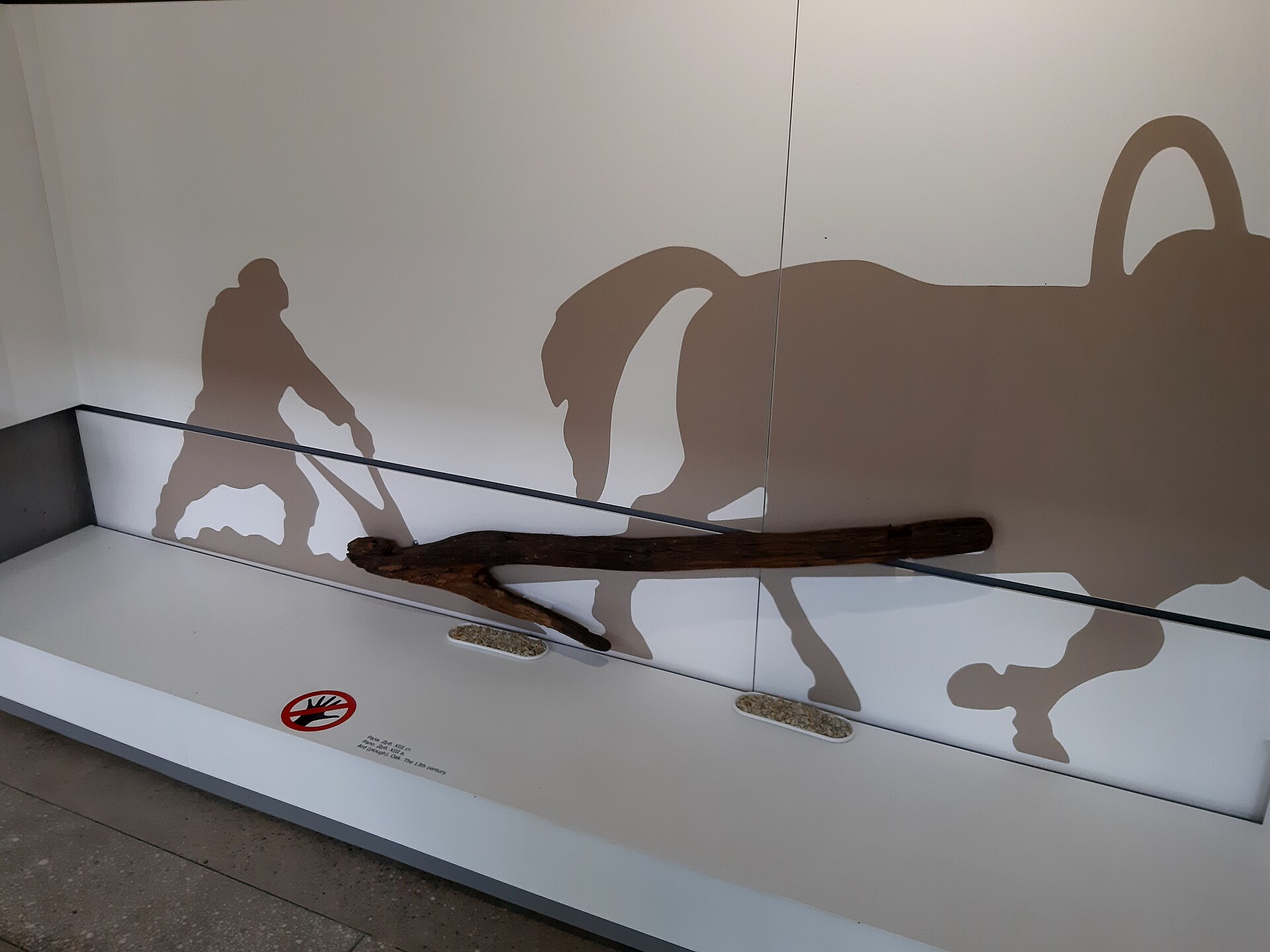 A silhouette panel of a man ploughing behind a horse, mounted above the museum's excavated wooden ard displayed flat in a glass case