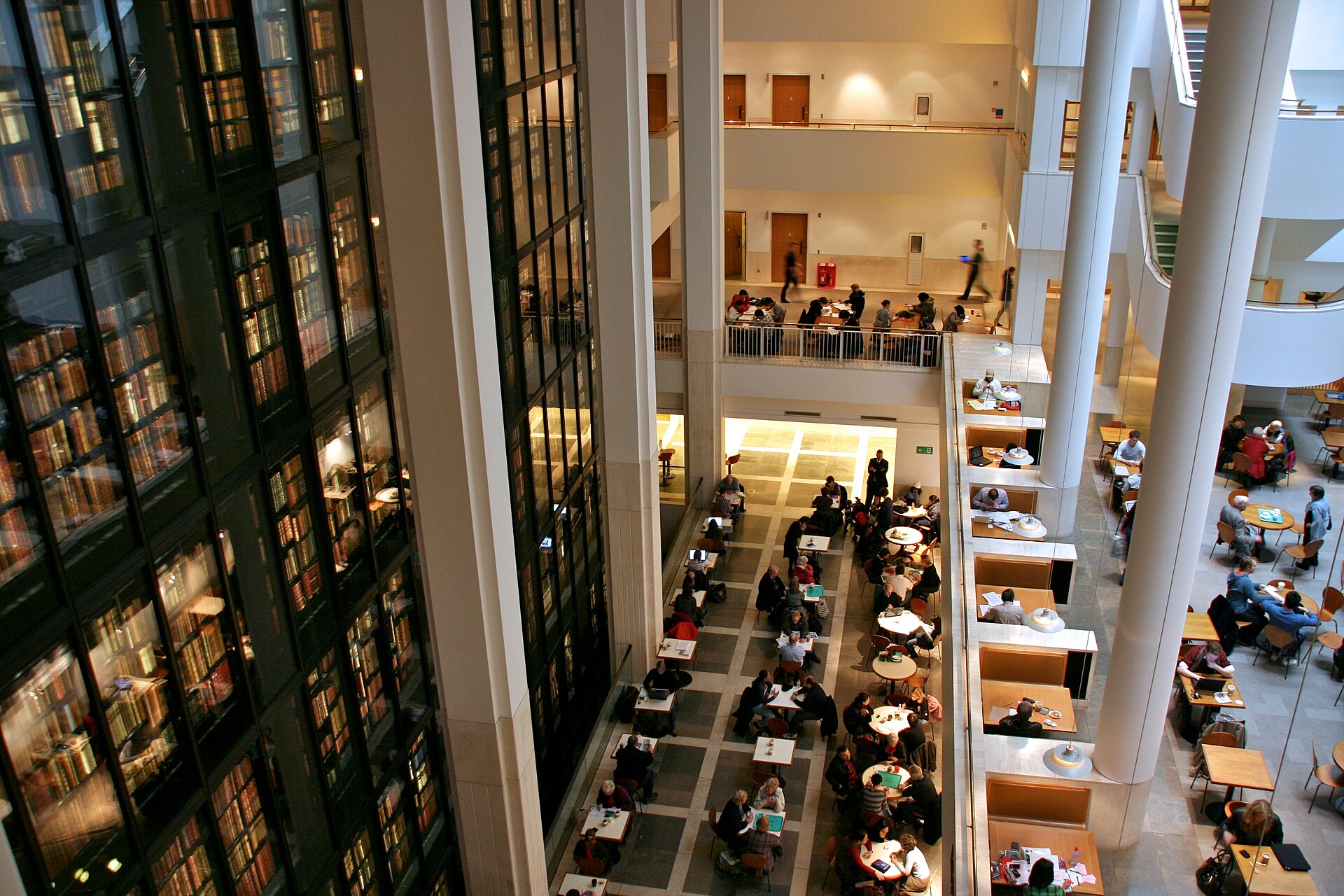 Reading room tables in the foreground with the tiered, black-shelved glass tower of the King's Library rising through several floors behind