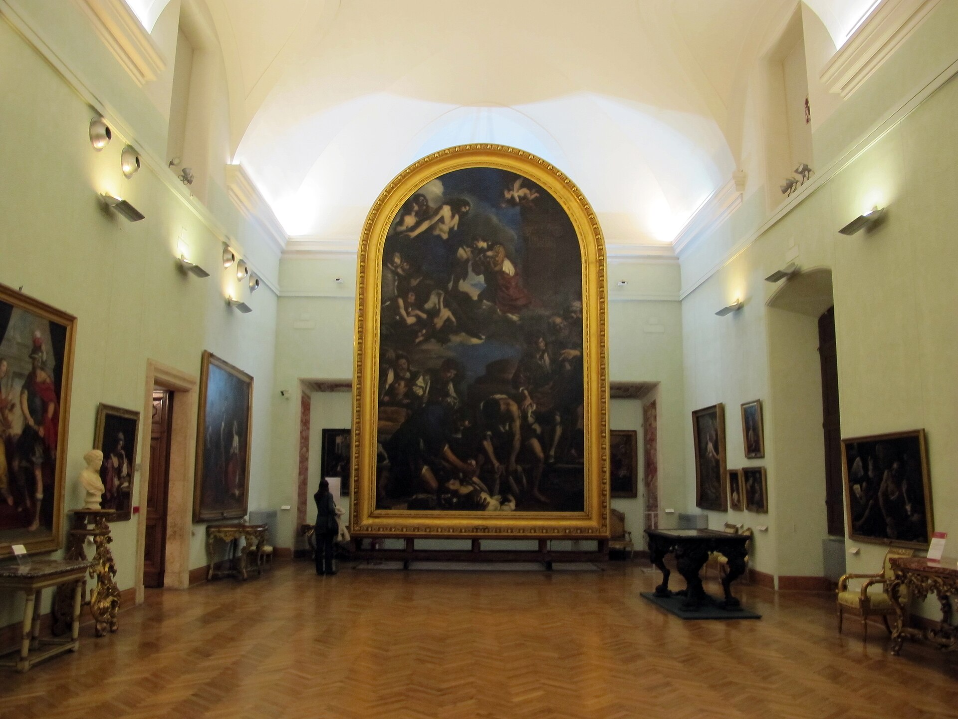 A high-ceilinged gallery with a very large altarpiece painting in a gilded arched frame, flanked by smaller portraits