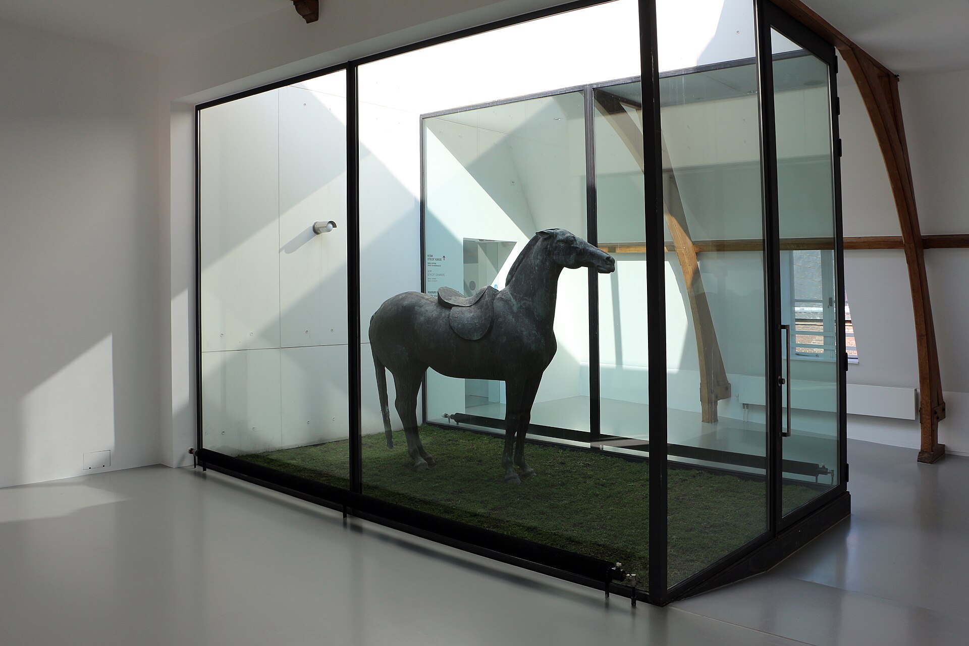 A dark sculpture of a horse in a glass display case within an attic gallery, exposed timber roof trusses visible above