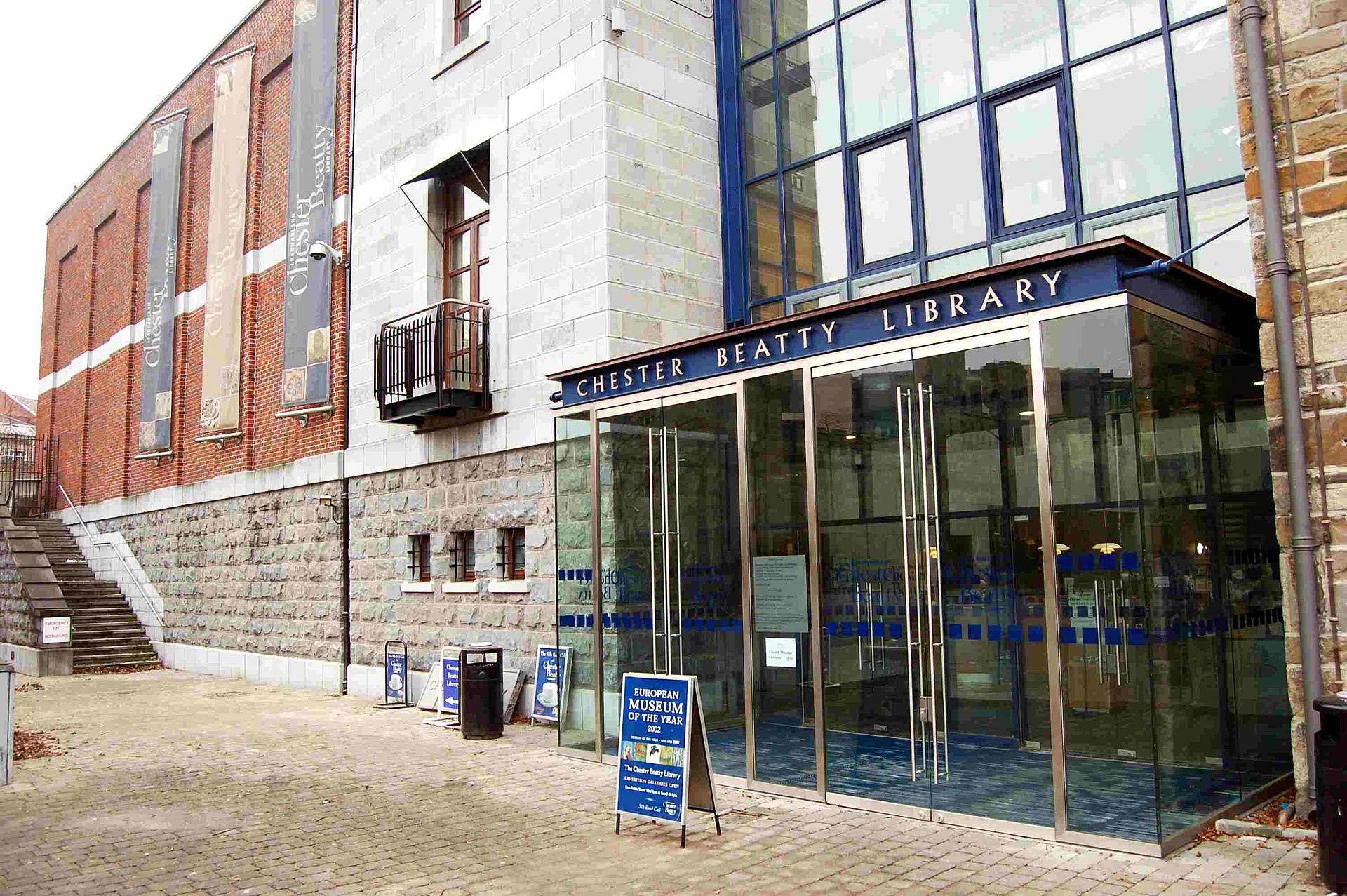 The stone and brick facade of the historic Clock Tower Building beside the glazed Chester Beatty Library entrance, with an upper balcony and mullioned windows visible