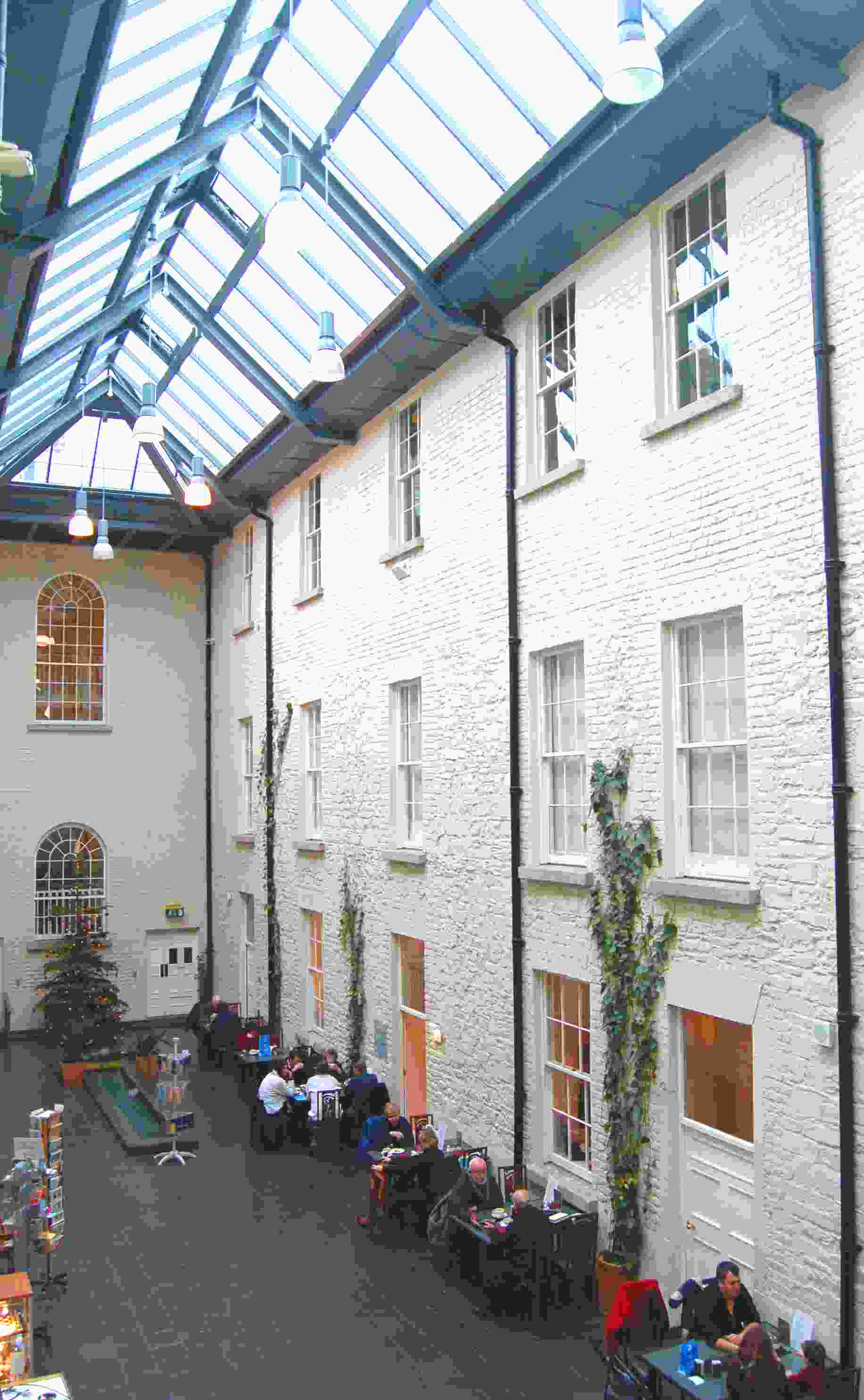 Looking down a long glazed atrium roofed in skylights, with cafe tables along a whitewashed wall below, linking the historic building to the exhibition wing