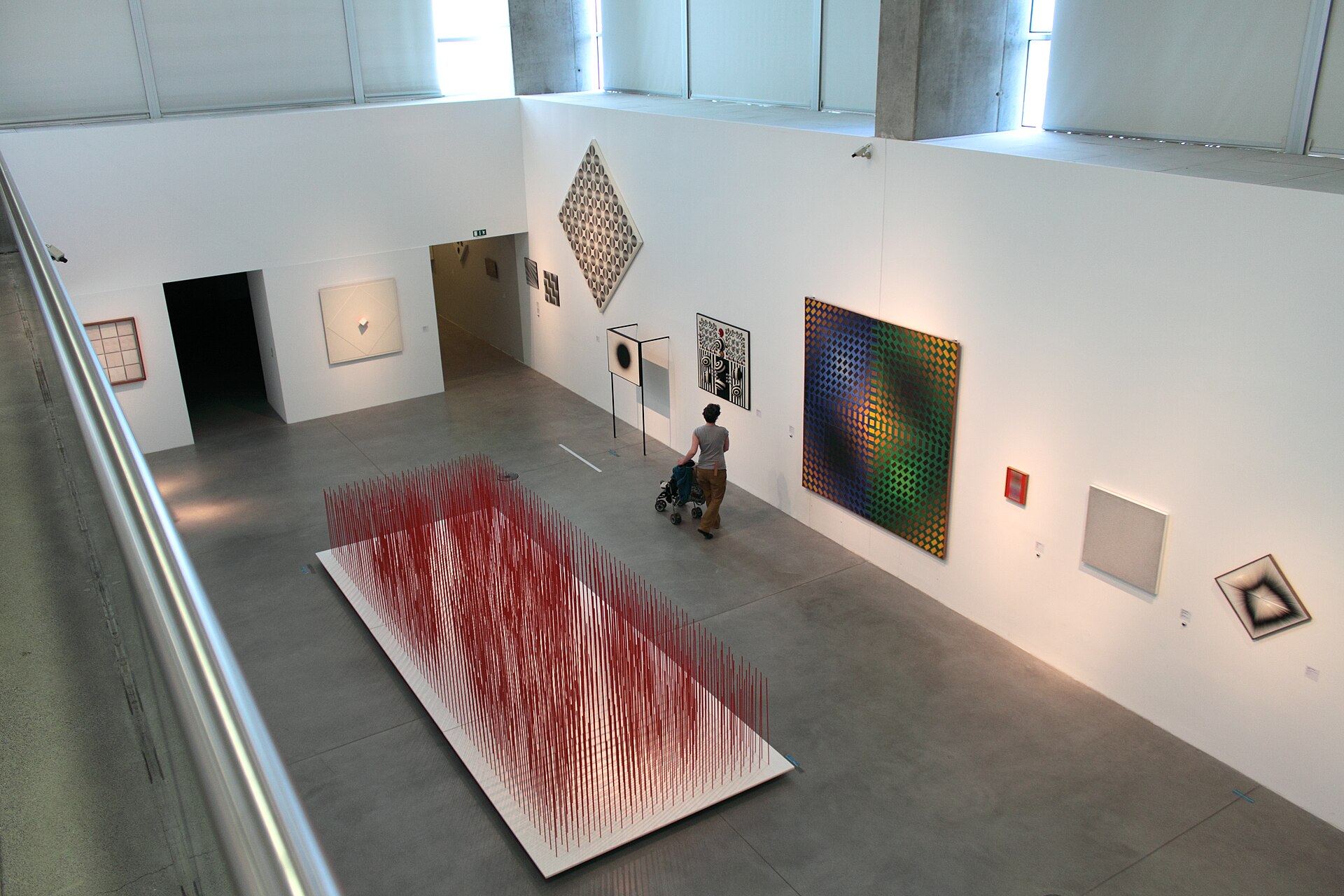 An elevated view into a double-height gallery hung with geometric abstraction paintings, including an optical diamond-shaped canvas, above a floor installation of vertical red strings
