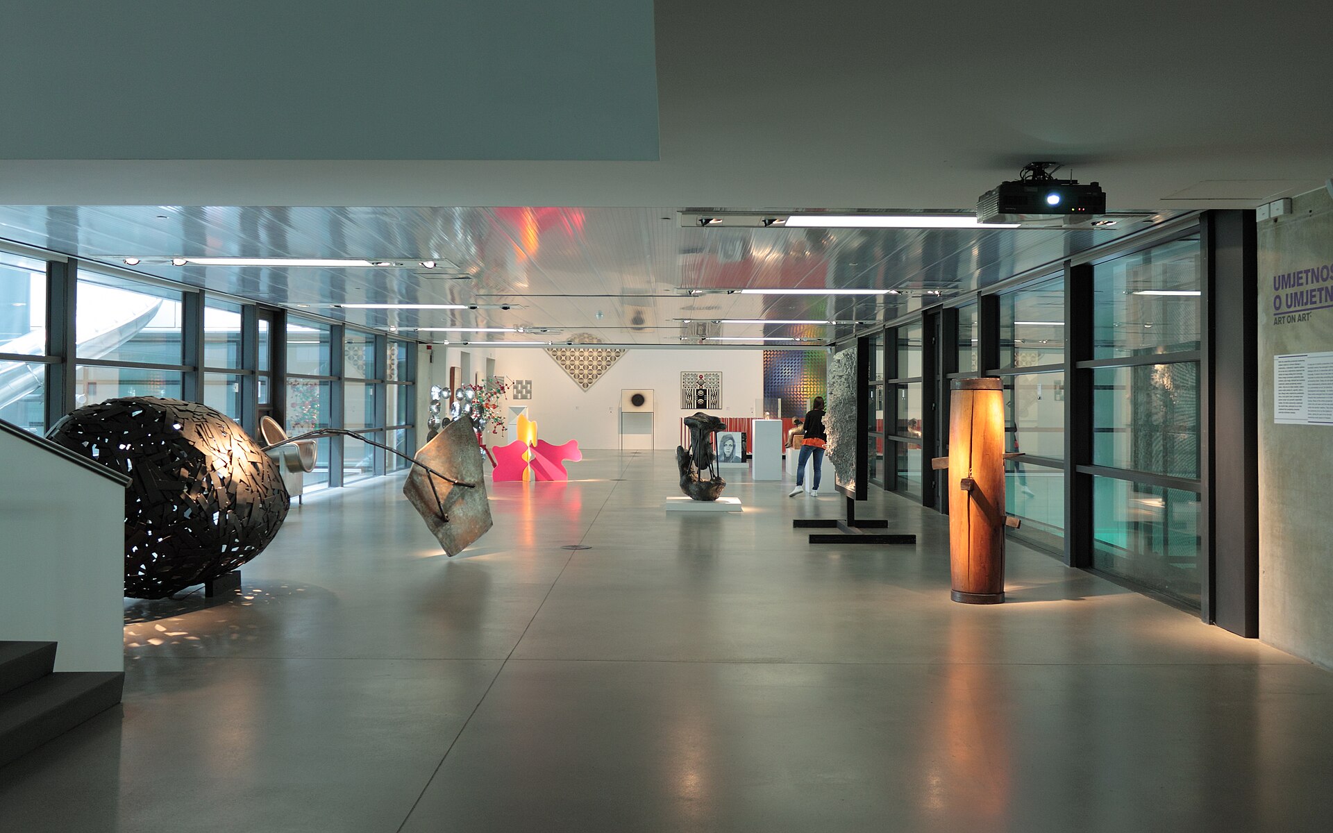 A corridor gallery of freestanding sculptural objects, including a dark animal-like form and a tall, lit orange column, next to a glass wall