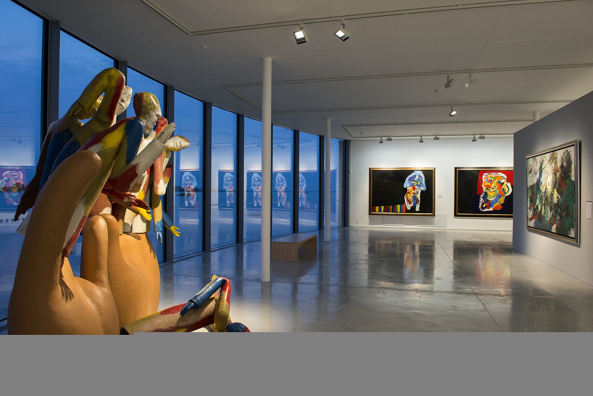 The curved, glass-walled extension pavilion at dusk, with a group of painted figurative sculptures in the foreground and paintings on the end wall.