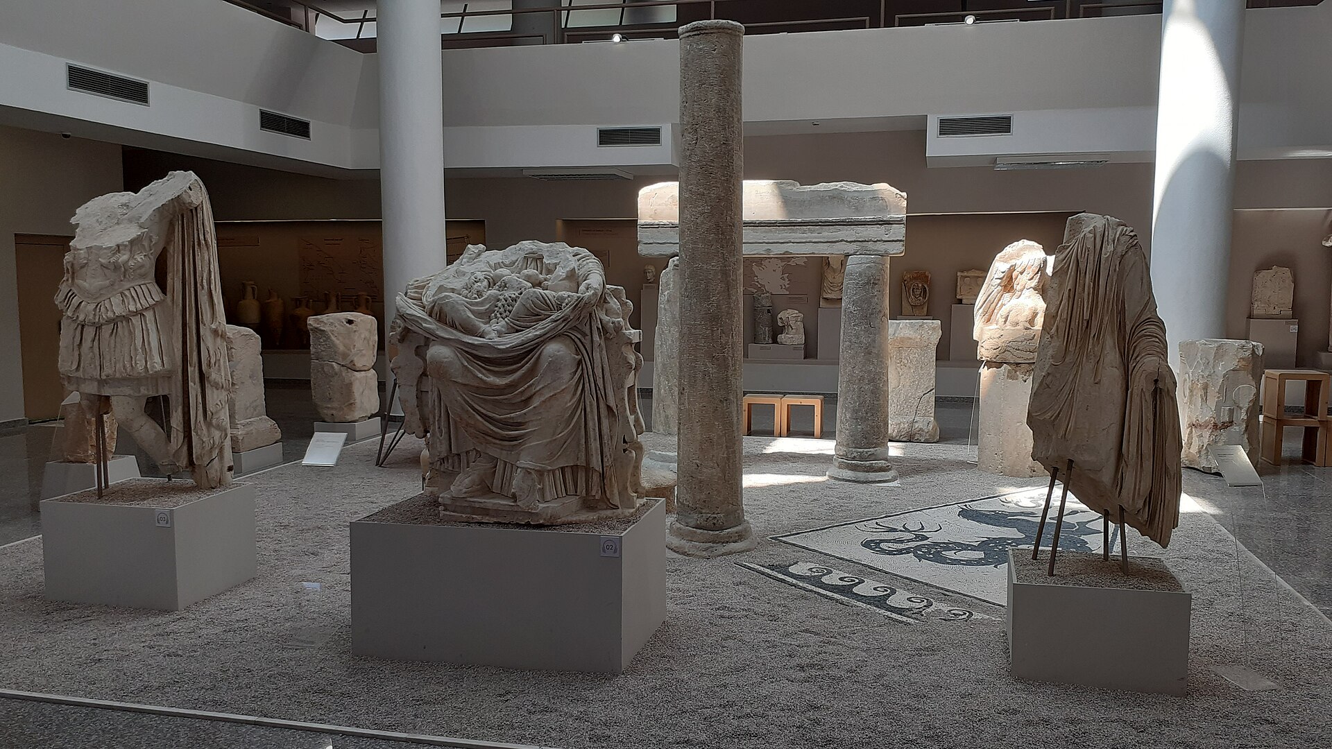 Central hall with large Roman marble statues arranged around ancient columns and a mosaic floor panel