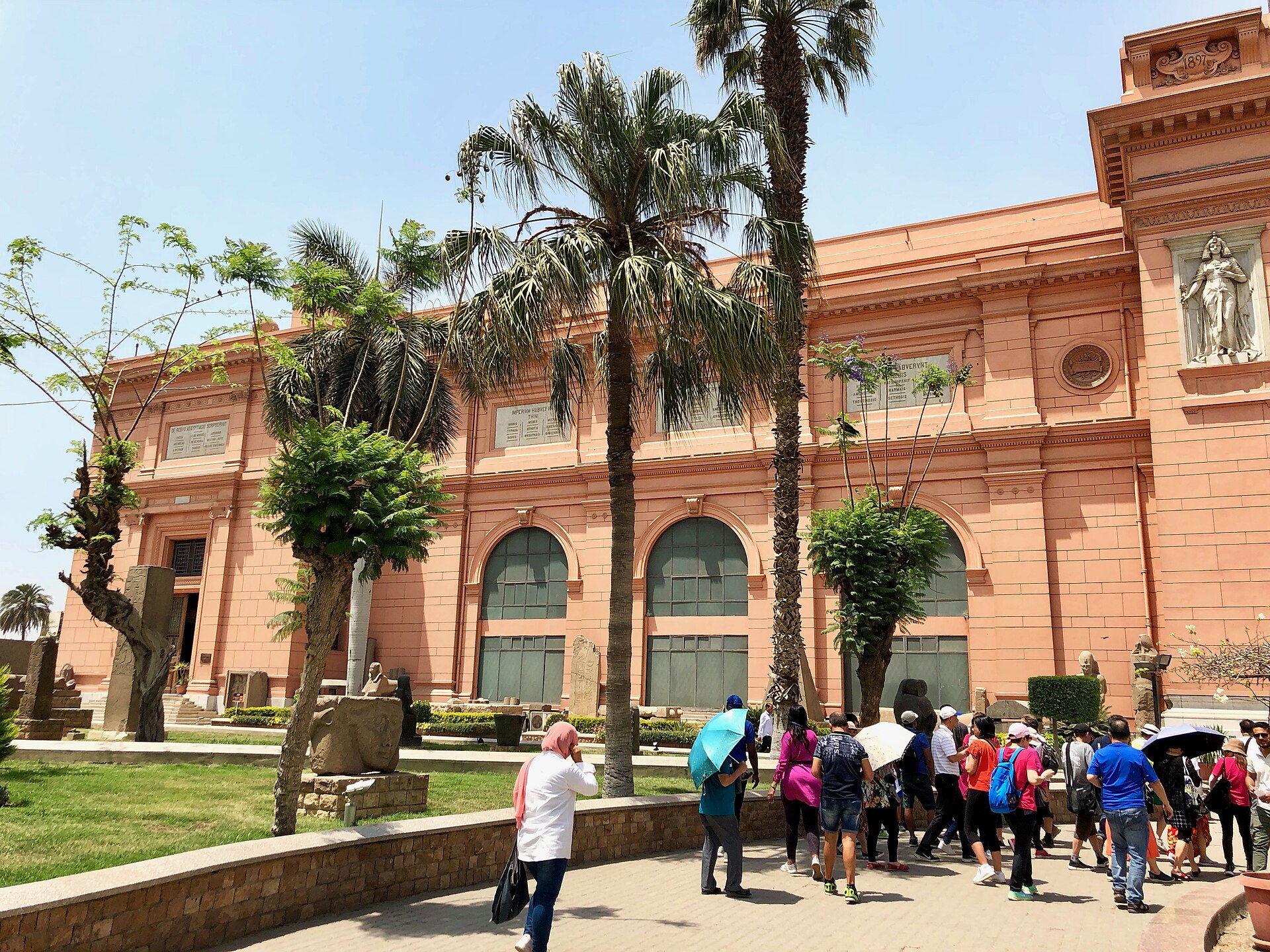 The pink facade seen from the garden, with palms, stelae set on the grass and the 1897 date on the end pavilion