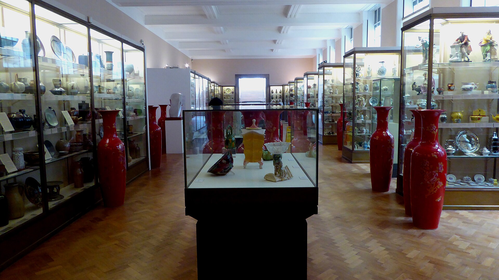 The Ceramics Gallery on the ground floor, a long parquet-floored room lined with glass display cases of European and Asian pottery and porcelain, with two tall red Chinese vases either side of a central low case