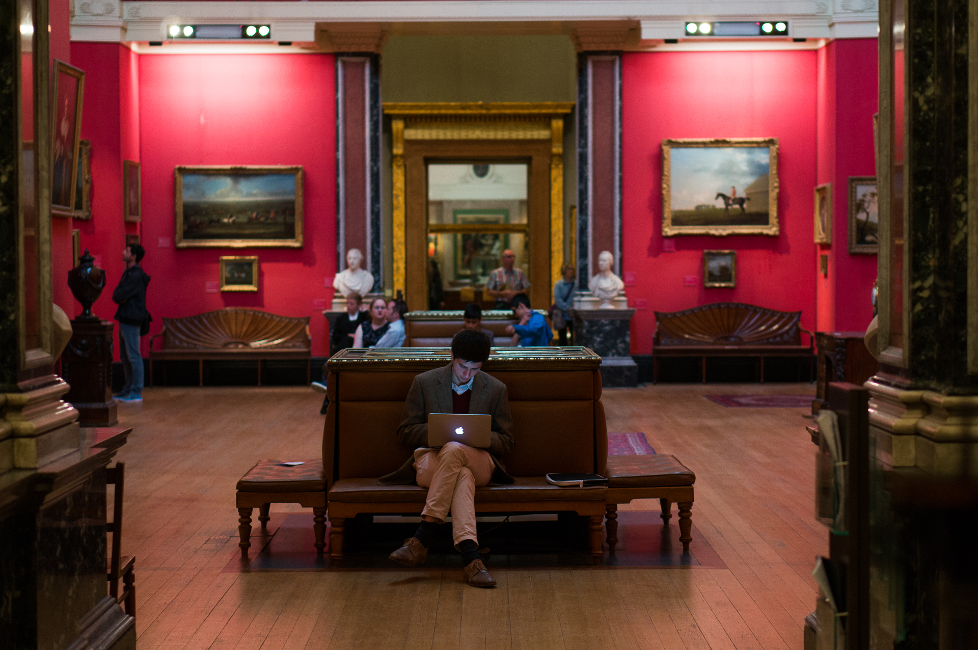 A red-walled Old Master picture gallery on the first floor, hung salon-style with gilt-framed paintings, with visitors seated on upholstered benches and marble busts on plinths against the wall