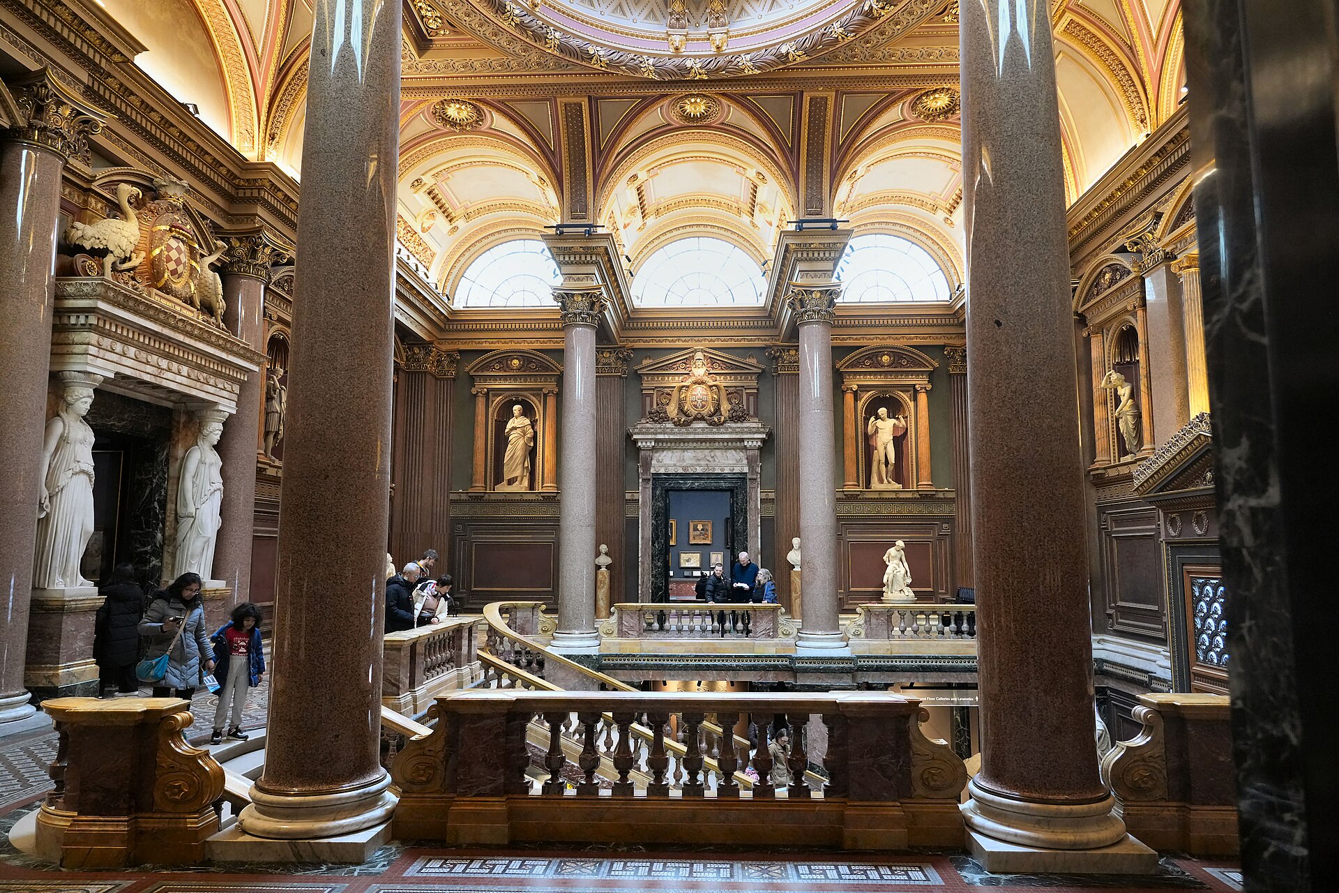 The Founder's Entrance Hall seen from the first-floor landing, showing the marble balustraded staircase, paired granite columns, niches with classical statues, and the coffered and domed ceiling above