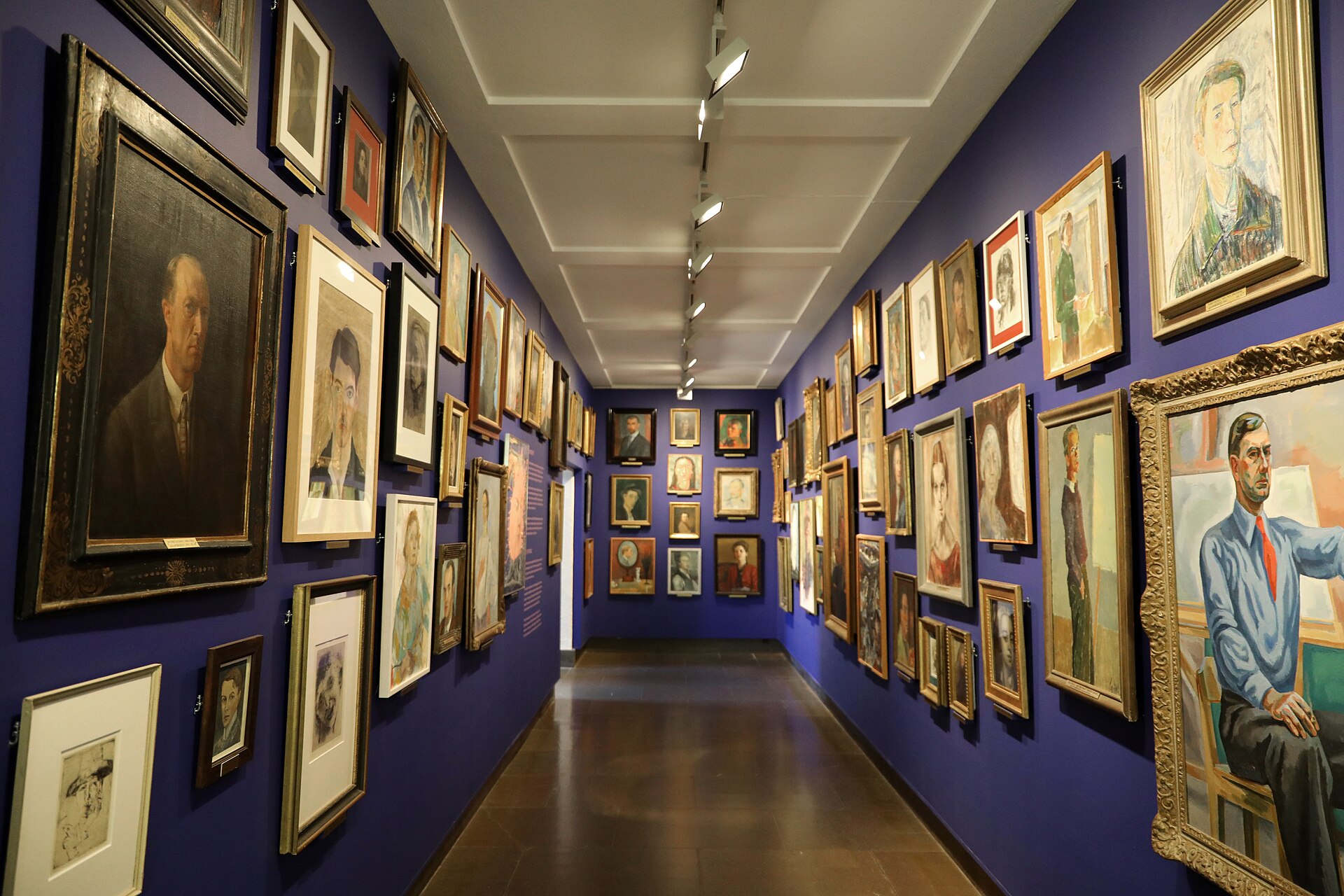 A long corridor gallery with dozens of self-portraits densely hung on dark blue walls