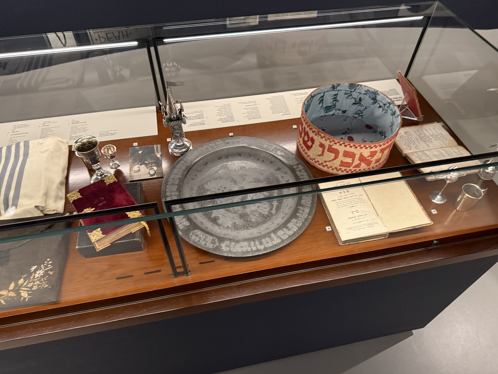 A museum display case of Judaica brought by German-speaking Jewish immigrants in the 1930s, including a Kiddush cup, an engraved Passover seder plate, prayer books and an embroidered cloth