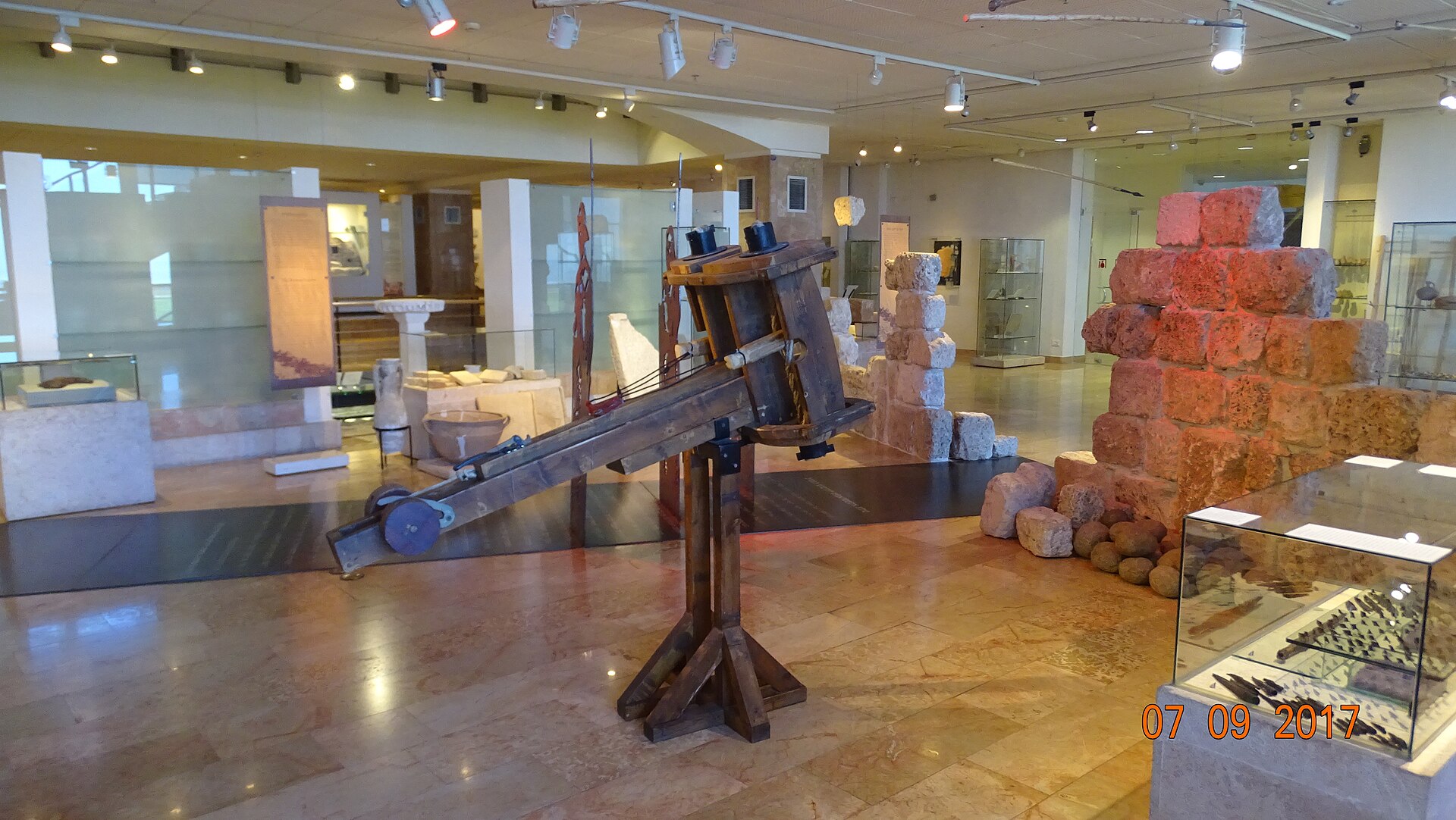 A two-level gallery hall in the Hecht Museum, seen from a staircase, with an Egyptian-style anthropoid coffin in the foreground and glass display cases of small finds along an upper balcony