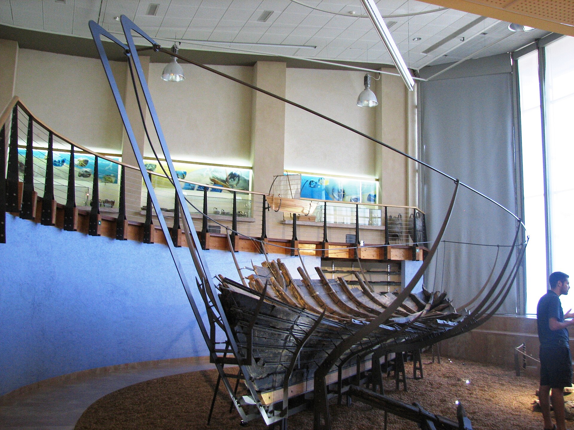 The reassembled wooden hull of the Ma'agan Michael Ship on its metal support cradle inside its dedicated museum wing, with explanatory panels on the curved wall behind it