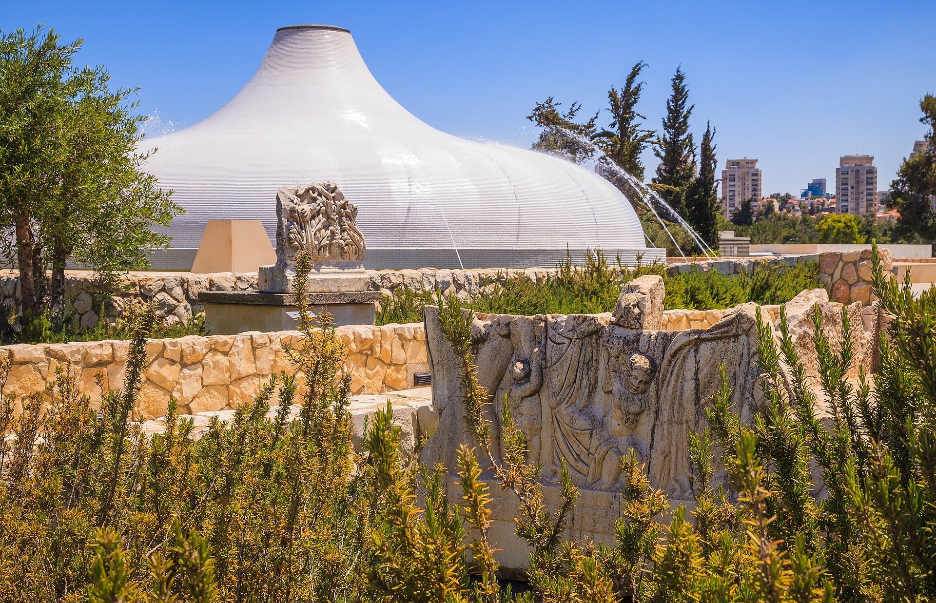 Ancient carved stone fragments displayed among rosemary bushes in the Billy Rose Art Garden, with the Shrine of the Book dome and its fountains behind