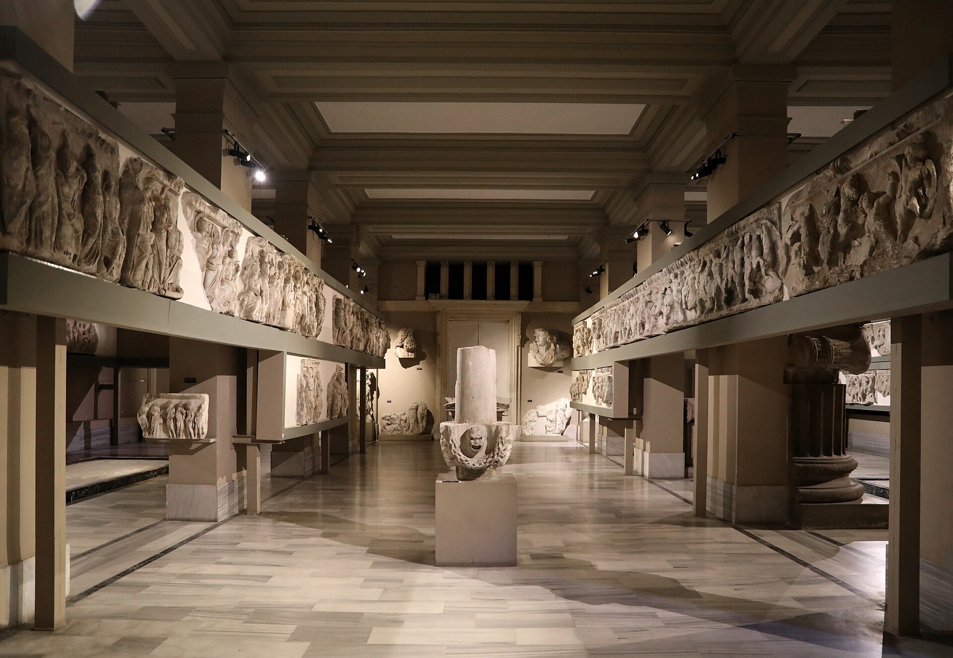 A long gallery with continuous carved marble friezes mounted high along both walls, an inscribed altar fragment standing at the centre of the marble floor