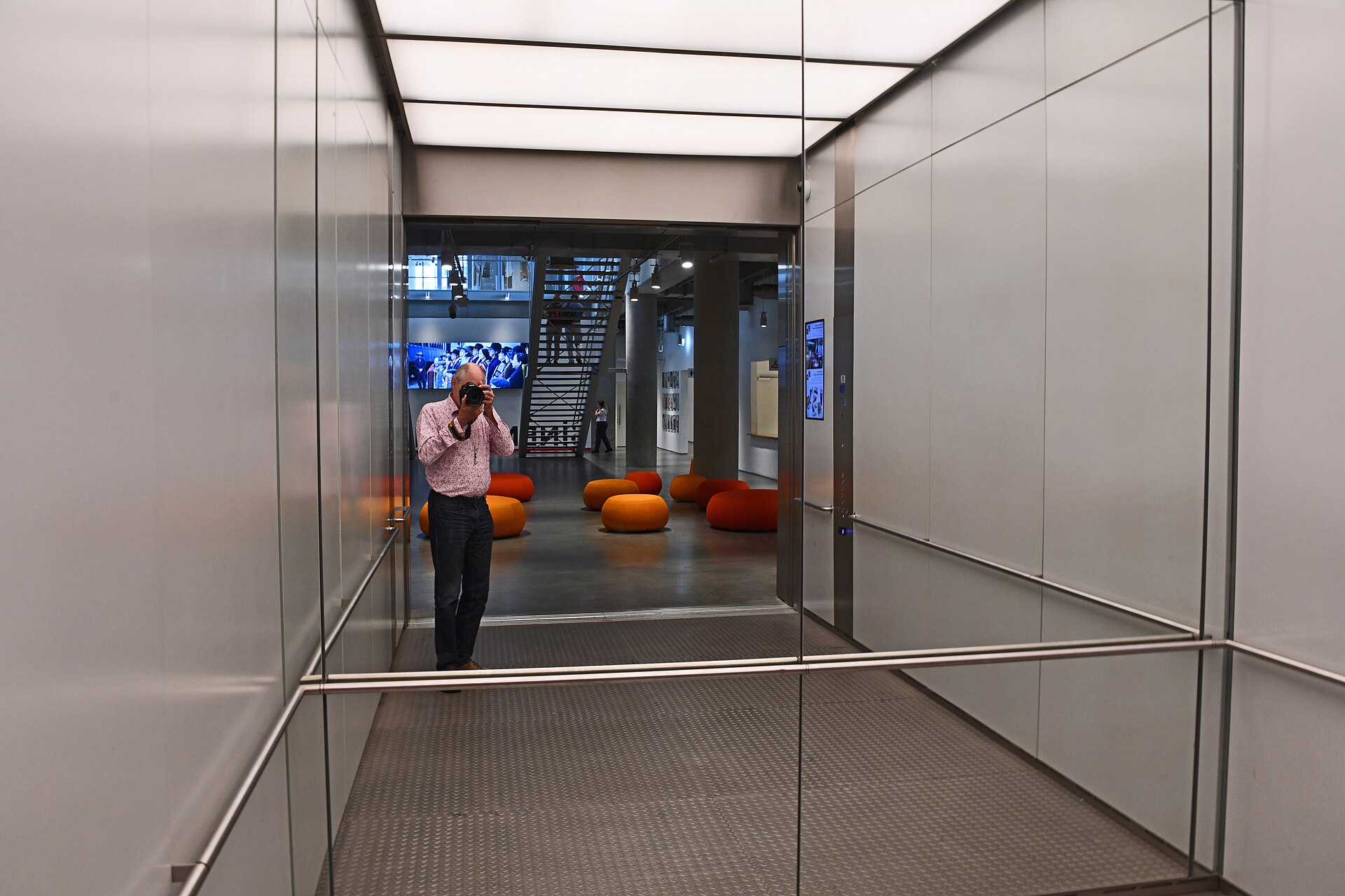 A glass-walled elevator and lobby area on the ground floor, with orange seating and museum signage visible beyond
