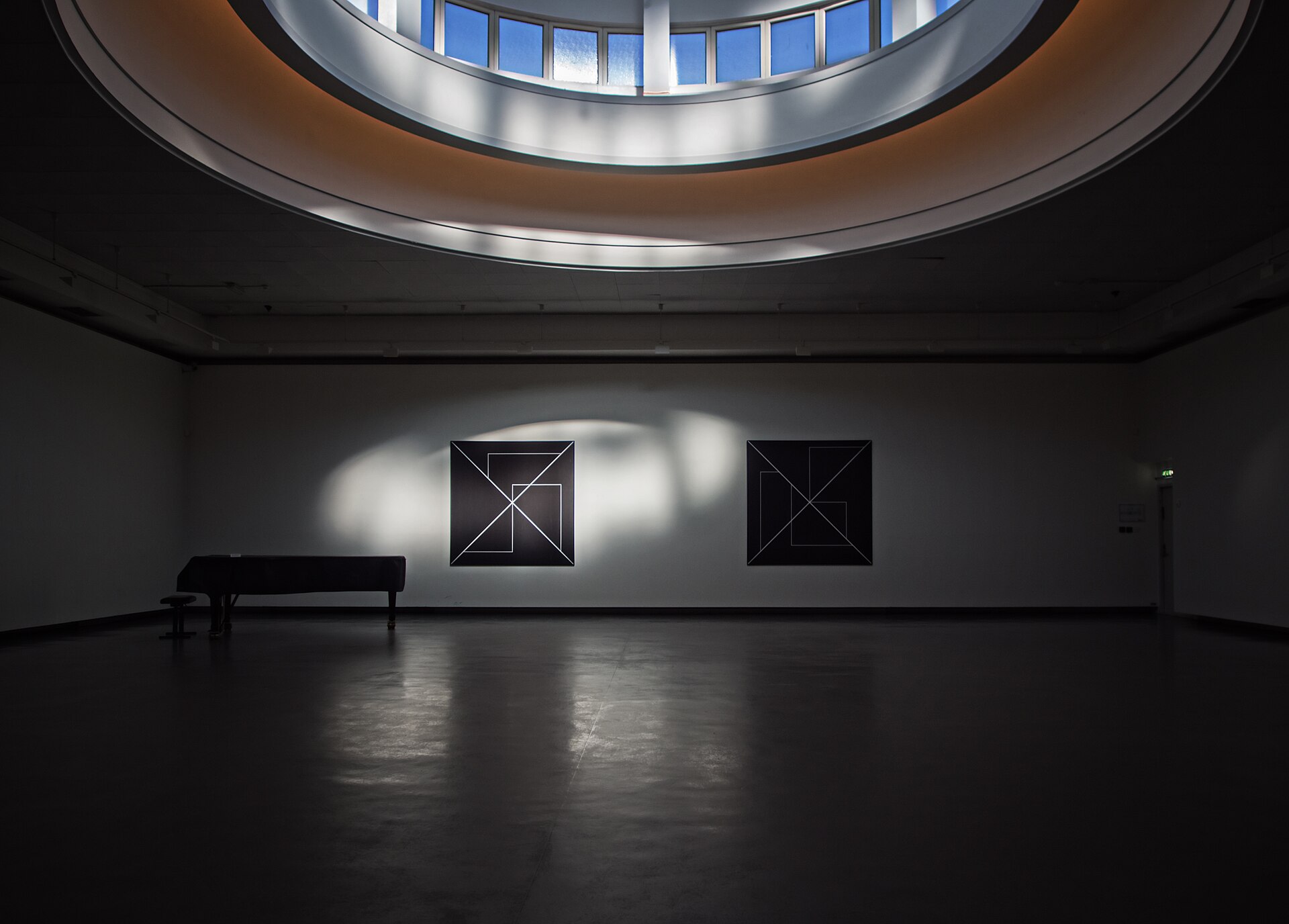A darkened gallery inside the circular Tarnsalen at Lysverket, with two geometric paintings lit by daylight from the ring of tower windows above and a grand piano in the foreground