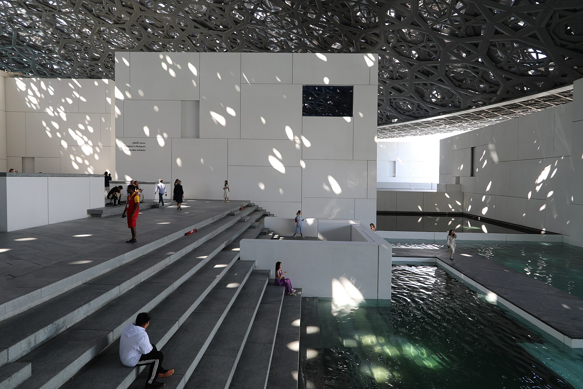 White concrete pavilion buildings and a water channel beneath the perforated dome, sunlight falling in a dappled pattern