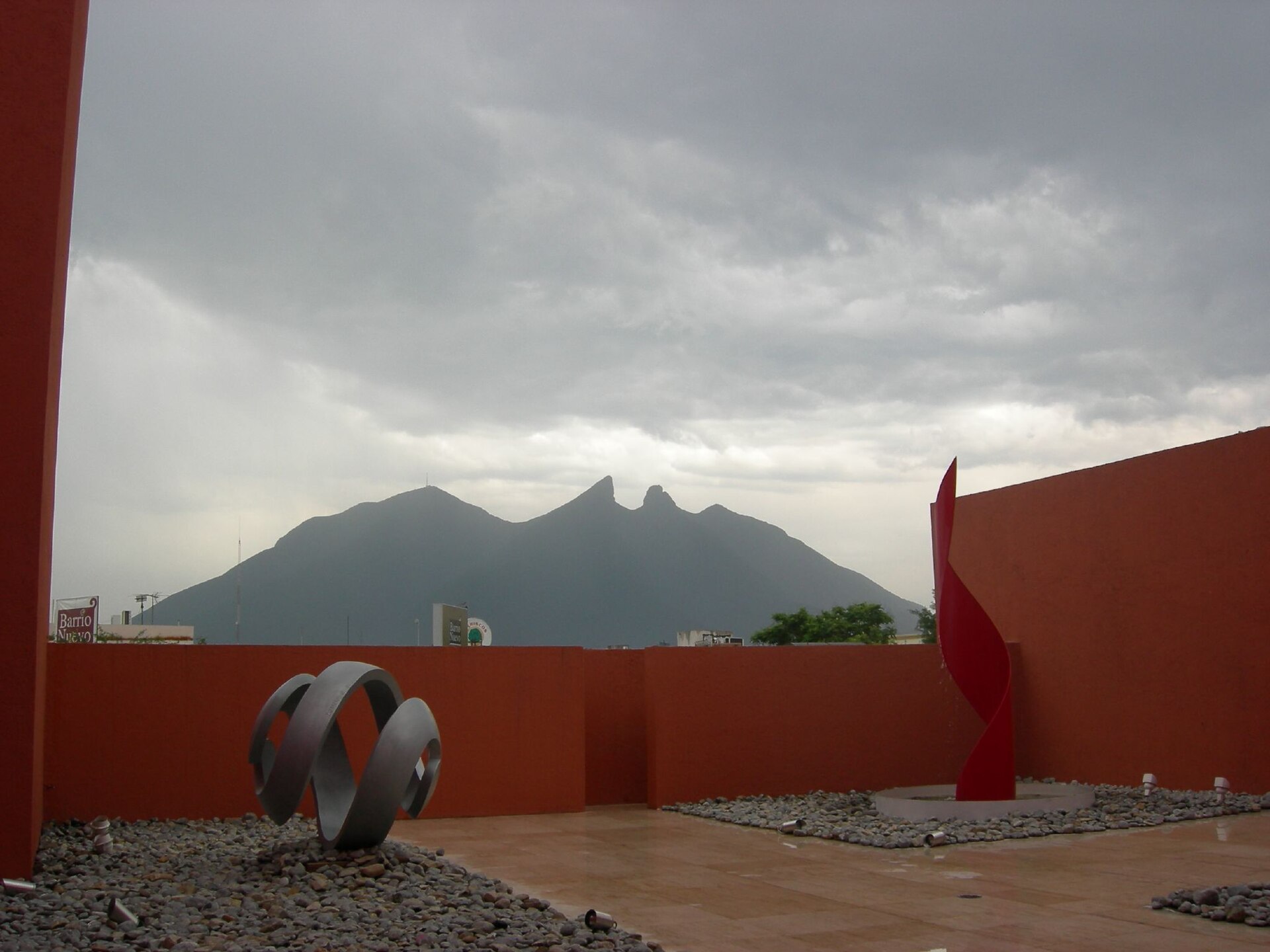 Two abstract sculptures on a terrace of river stone, with the peaks of the Cerro de la Silla visible above the terracotta perimeter wall