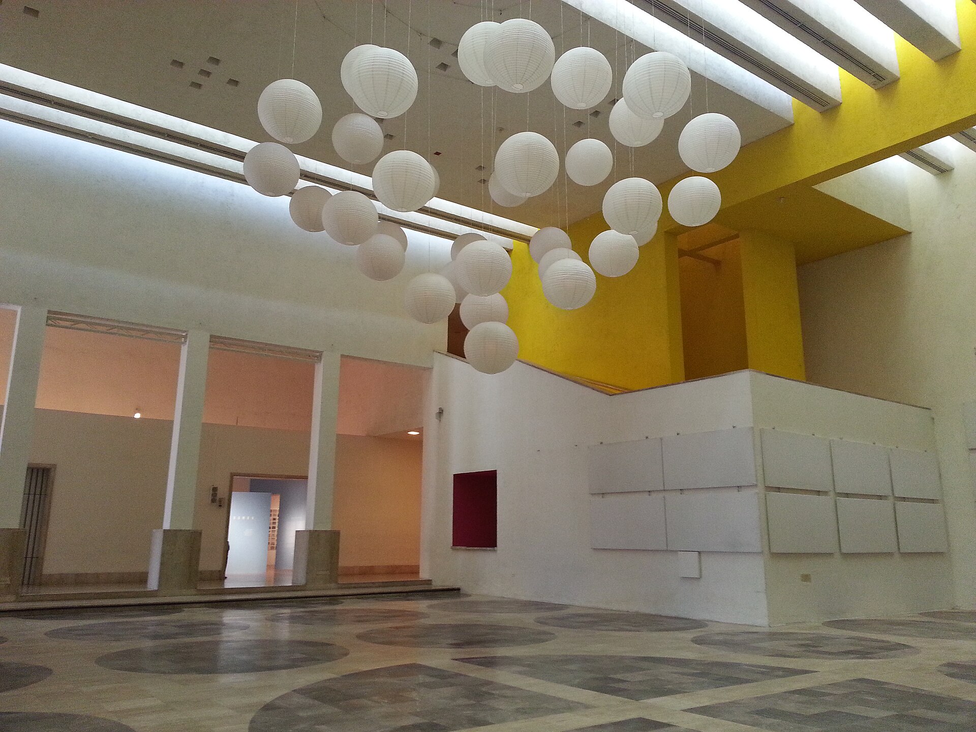 A high-ceilinged gallery hung with a suspended installation of round paper lanterns, with further exhibition rooms visible through doorways beyond