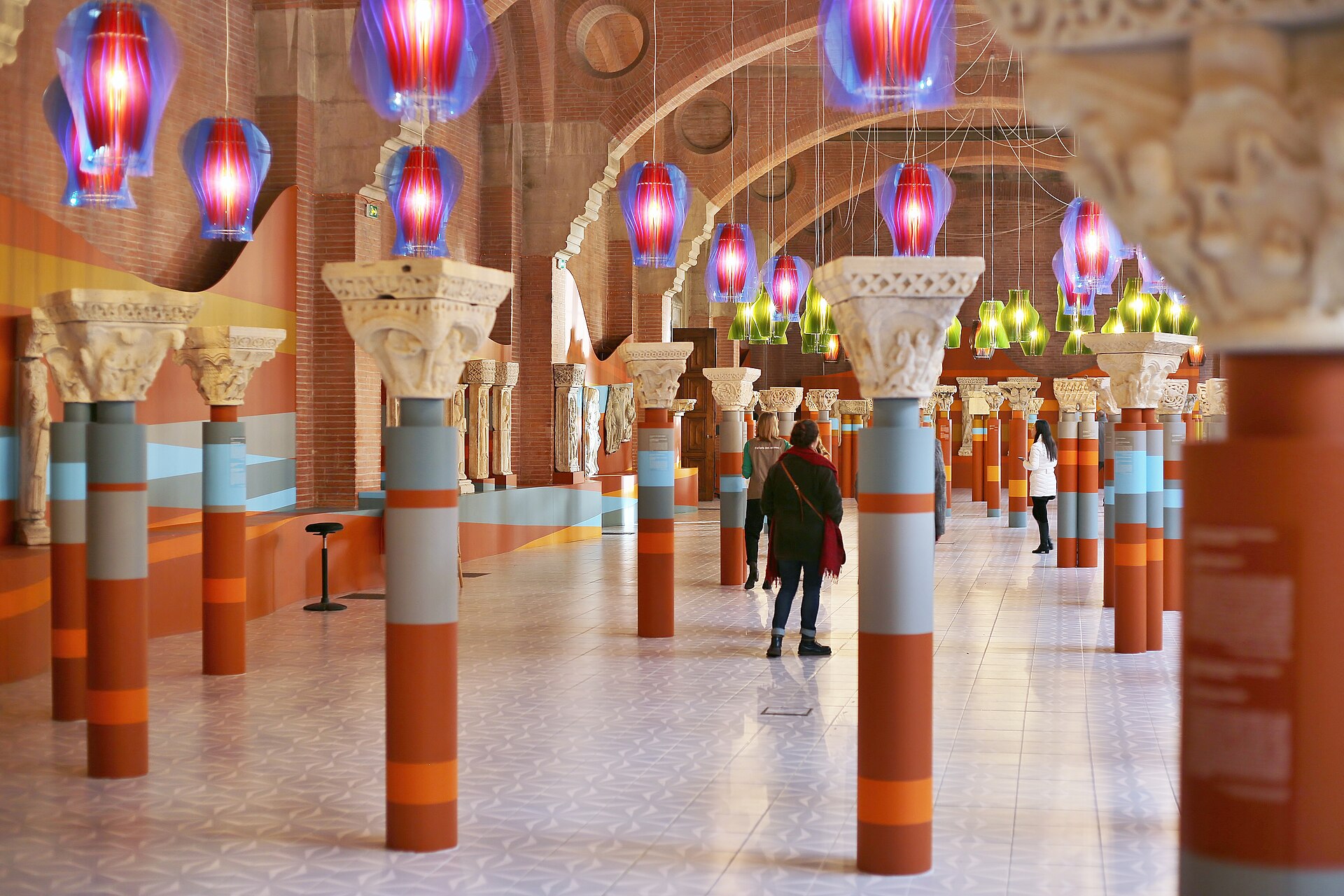 A vaulted brick hall lined with rows of carved Romanesque capitals on brightly painted red and blue columns, hung with colourful glass lamps, with visitors walking between them.