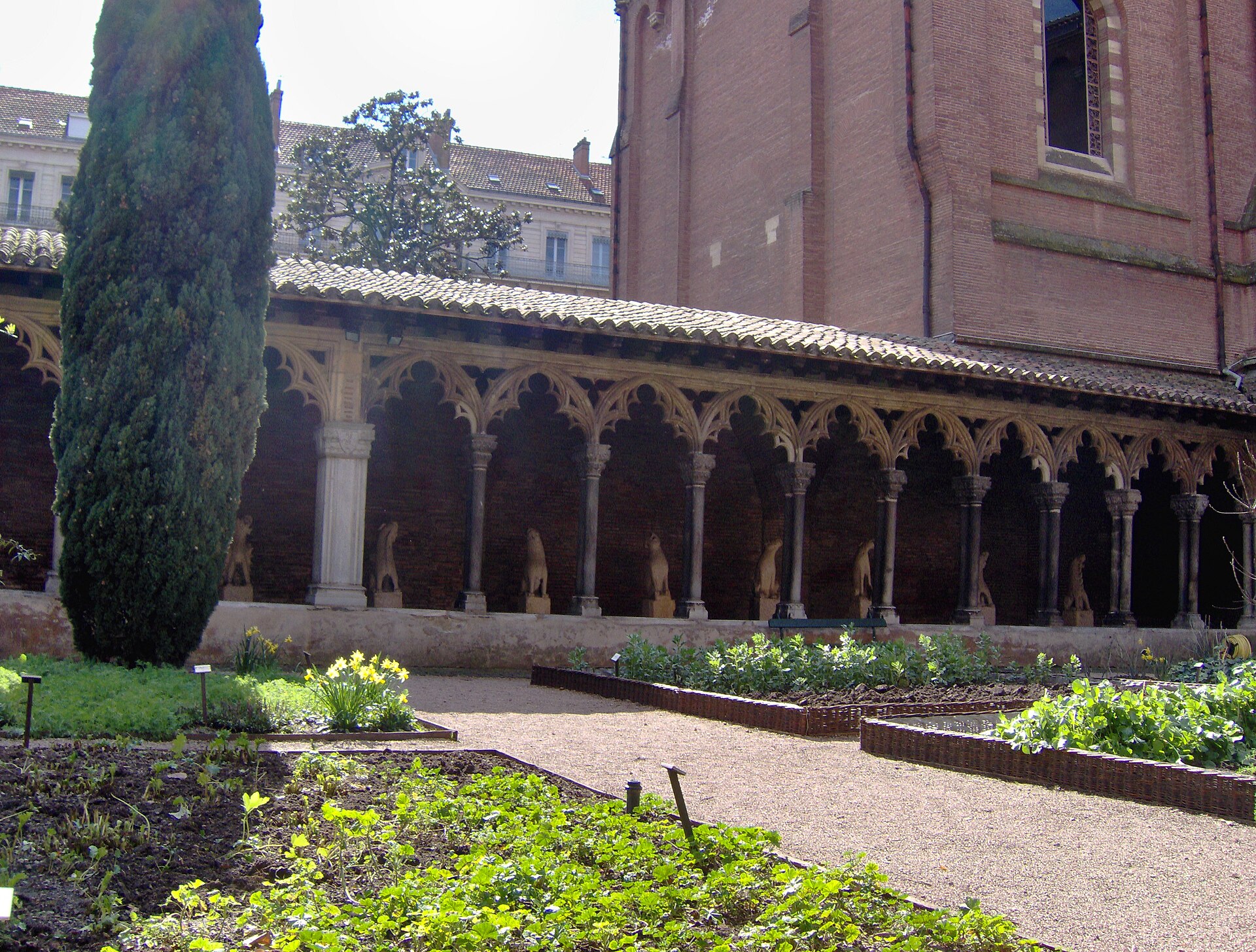 A brick-arcaded medieval cloister walk with a row of carved marble columns and figures under a tiled roof, seen across garden flowerbeds and a tall cypress tree.