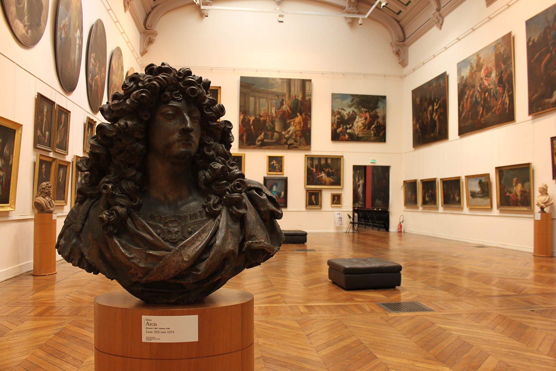 A tall salon with paintings hung salon-style floor to cornice on cream walls under a glazed roof, with a dark bronze portrait bust of Louis XIV on a plinth in the foreground.