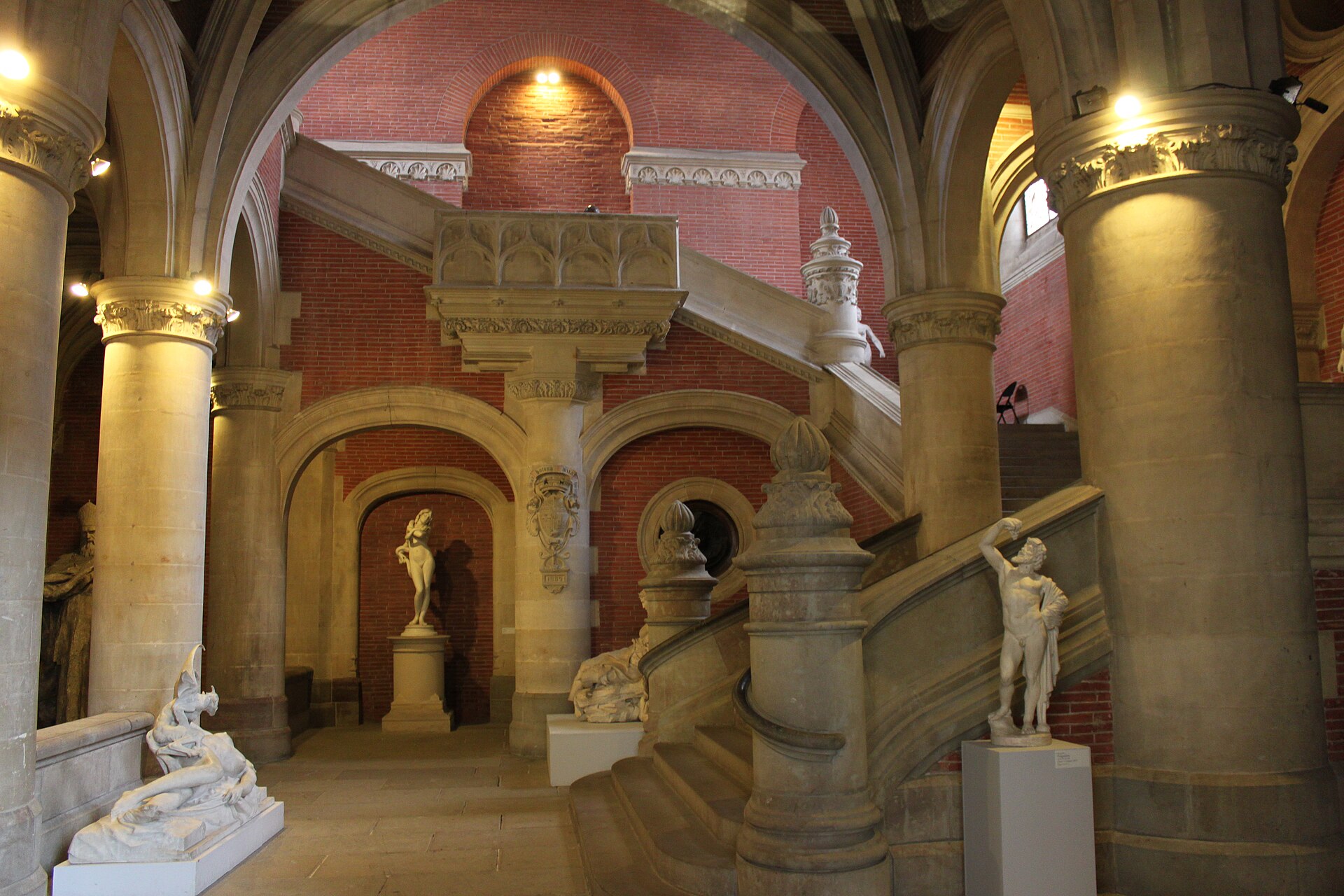 A grand stone staircase rising beneath red-brick arches, with white marble and bronze figure sculptures on plinths at its foot.