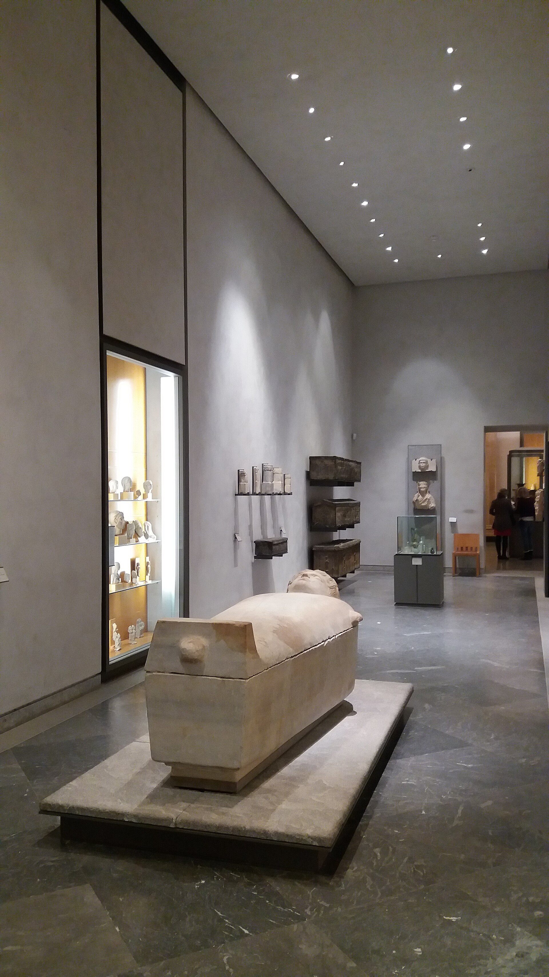 A gallery of Egyptian antiquities with a stone sarcophagus in the foreground and wall cases of smaller funerary objects.