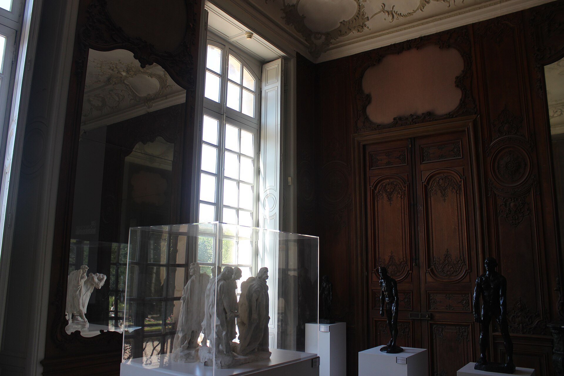 A panelled room in the historic house, with a tall window, an antique mirror and sculptures under glass