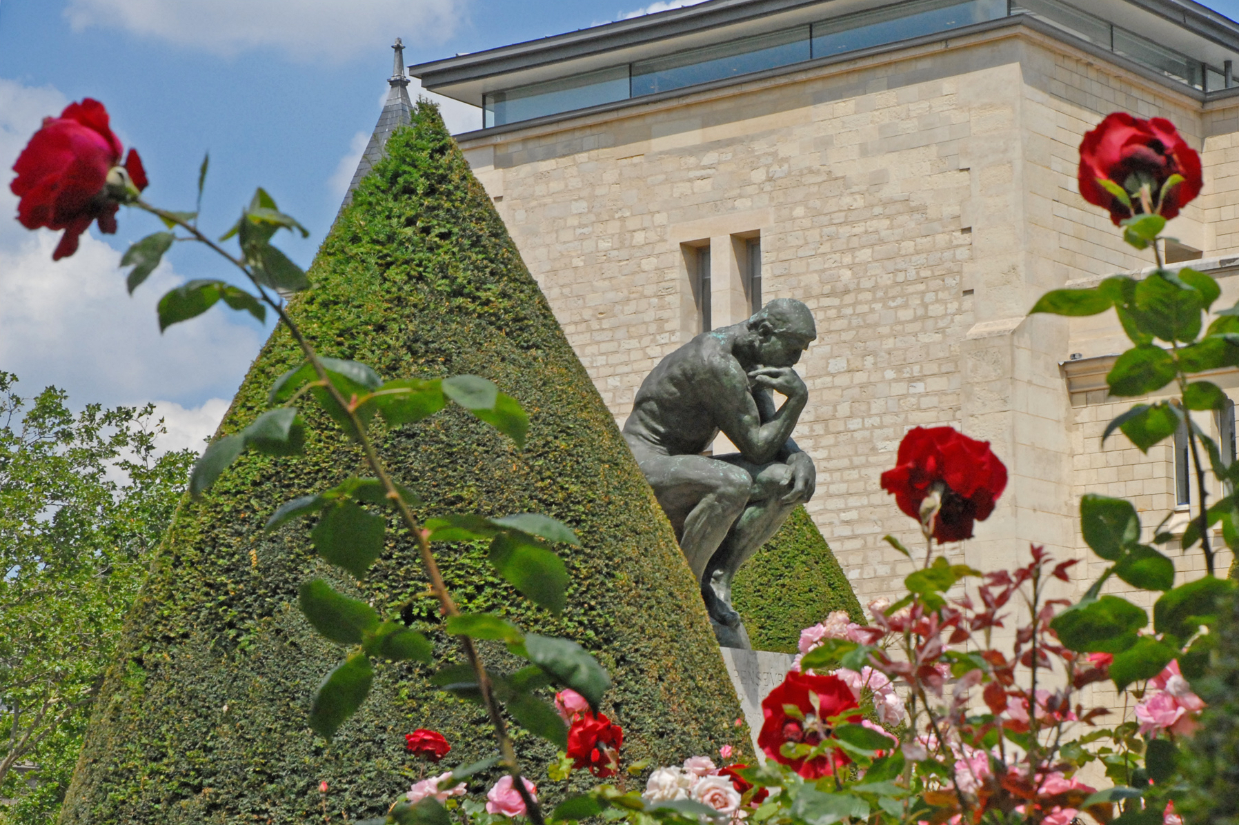 The monumental bronze Thinker among clipped hedges in the garden, with a corner of the museum building and roses in the foreground