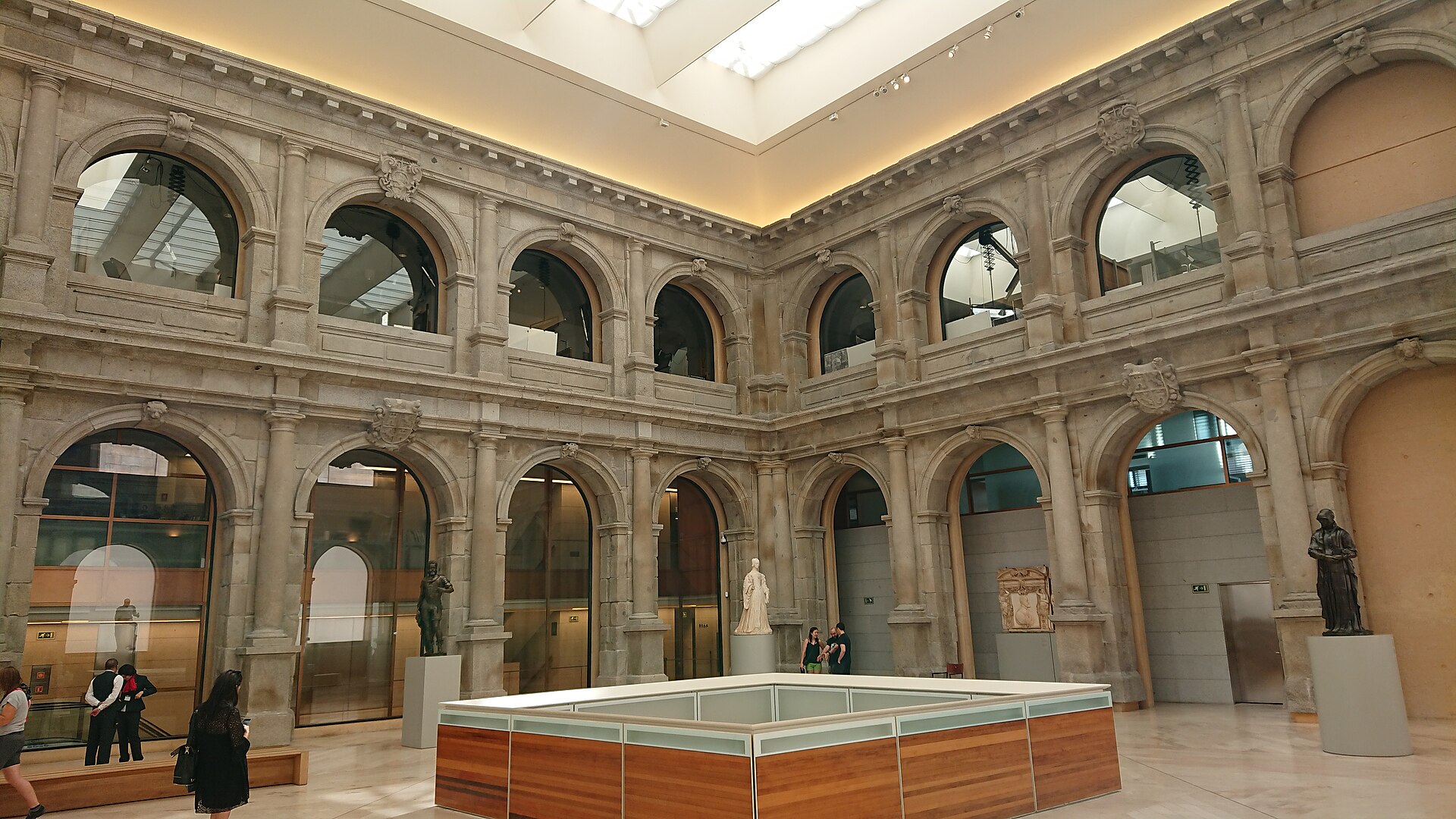 A two-tier granite cloister arcade glazed and enclosed under a top-lit ceiling, with sculpture on plinths on the floor
